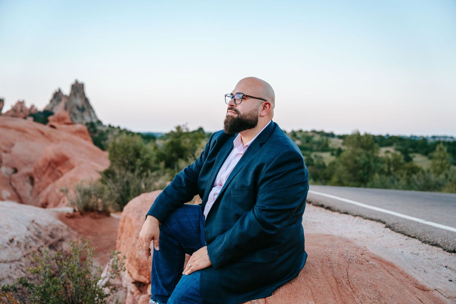 Bald bearded man in navy suit sitting thoughtfully on red rocks with desert landscape at dusk.