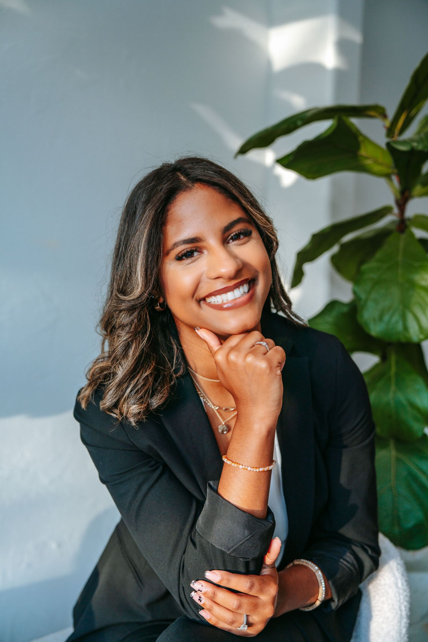 Joyful professional woman in black blazer laughing with hand on chin beside a lush green indoor plant.