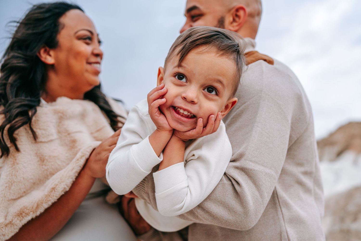Happy young child resting chin on hands smiling at camera with parents behind outdoors.