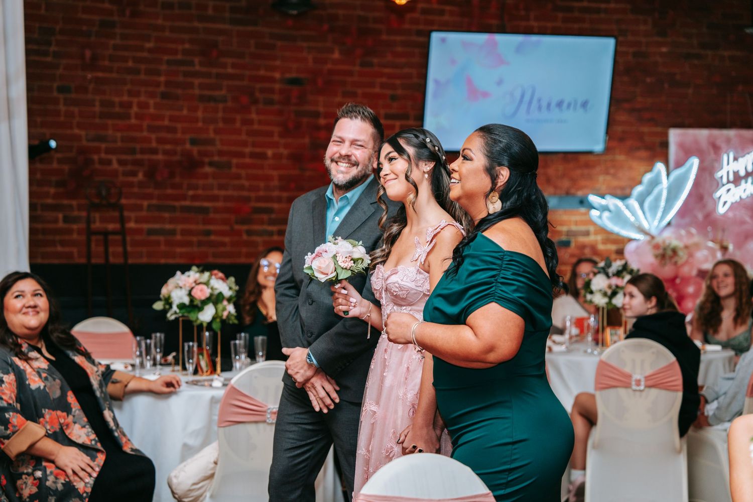 Quinceañera girl in pink gown escorted by parents into reception hall with decorative backdrop.