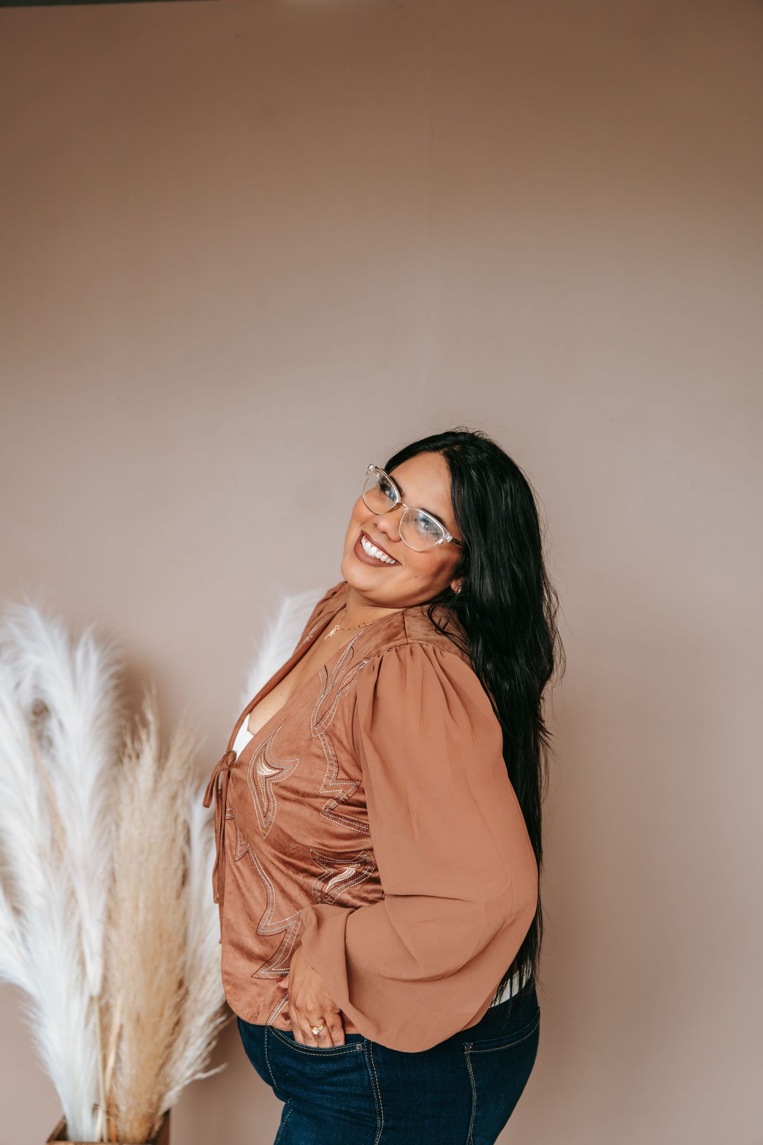 A woman with long dark hair and glasses smiles over her shoulder in a brown blouse beside pampas grass.