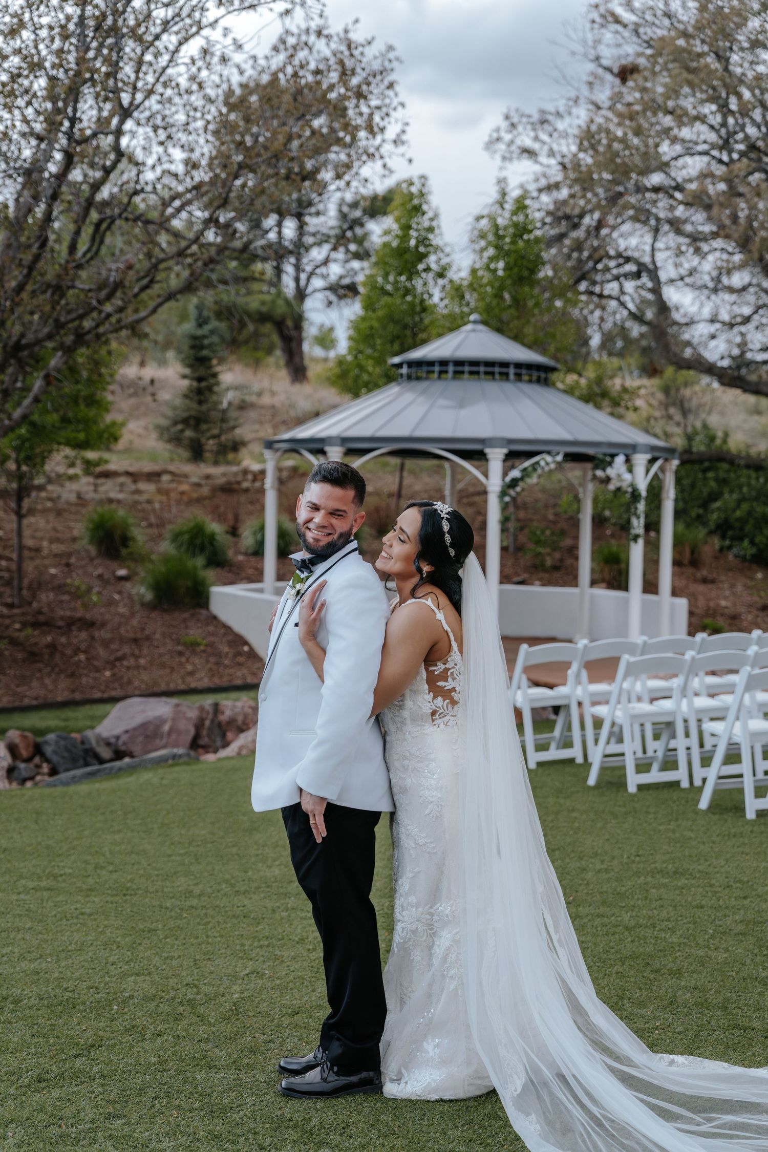 Bride and groom pose together in front of a white gazebo on a lush green lawn at their wedding.