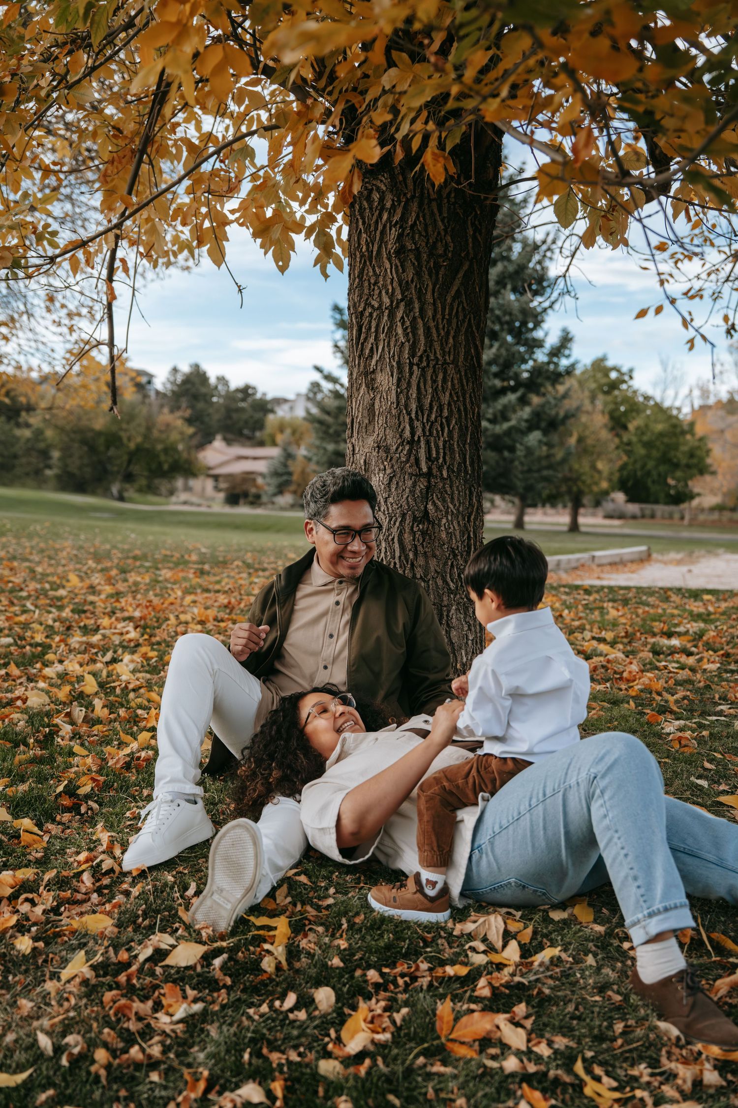 Family of three relaxing under a large tree surrounded by golden autumn leaves in a park.