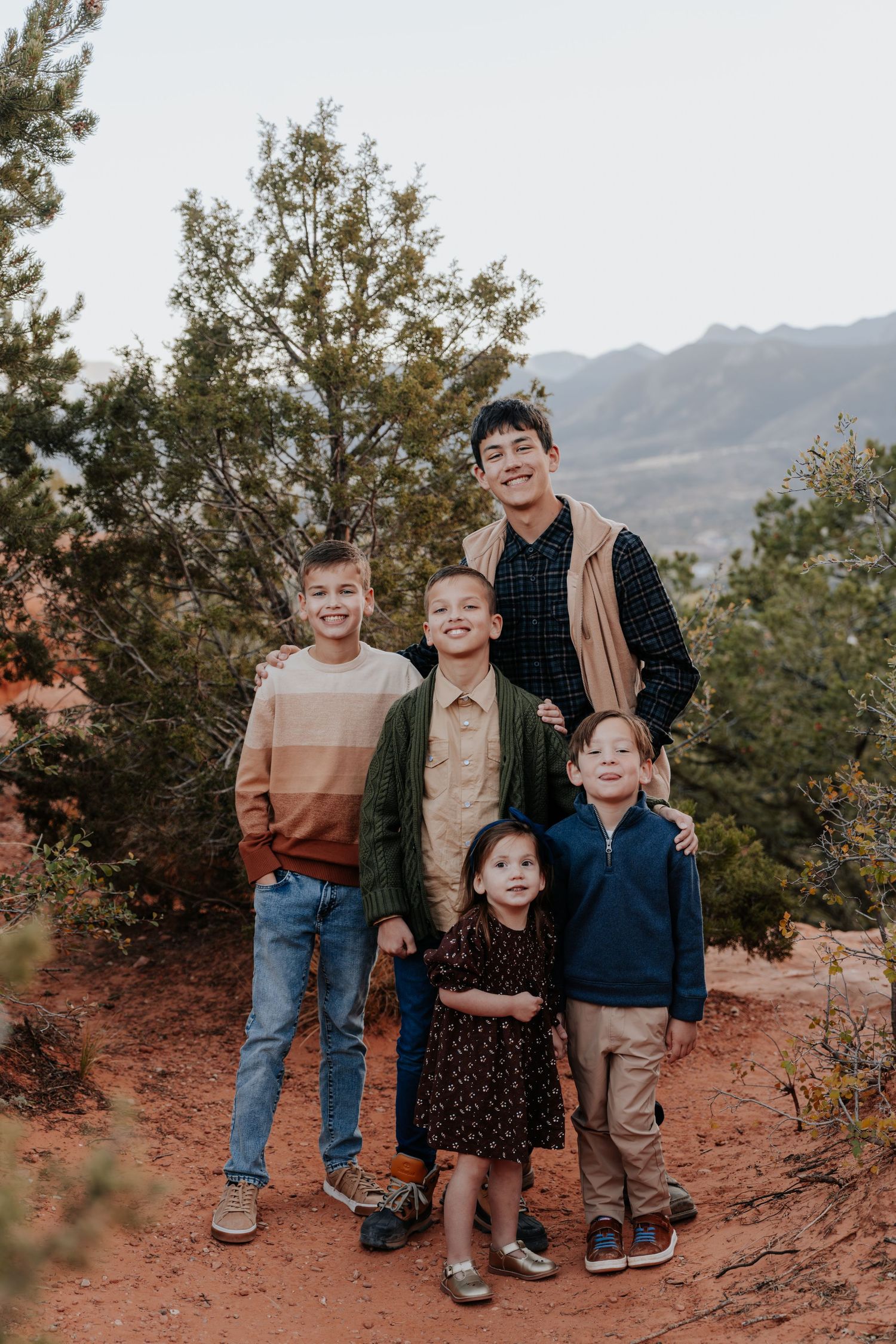 Five children posing together on red dirt trail surrounded by trees and mountain landscape.