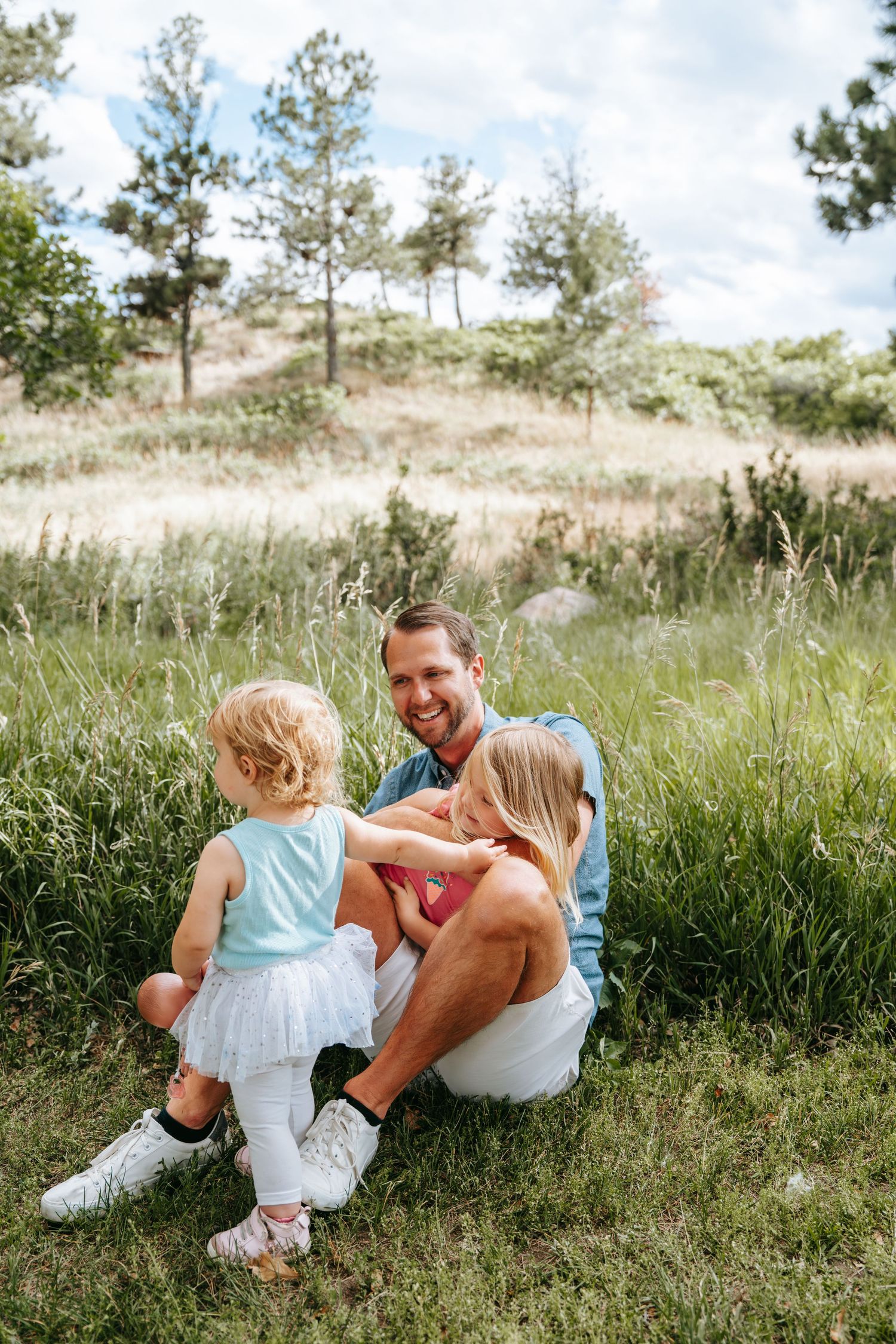 A smiling man sits on grass with two young girls in a lush green outdoor setting on a cloudy day.