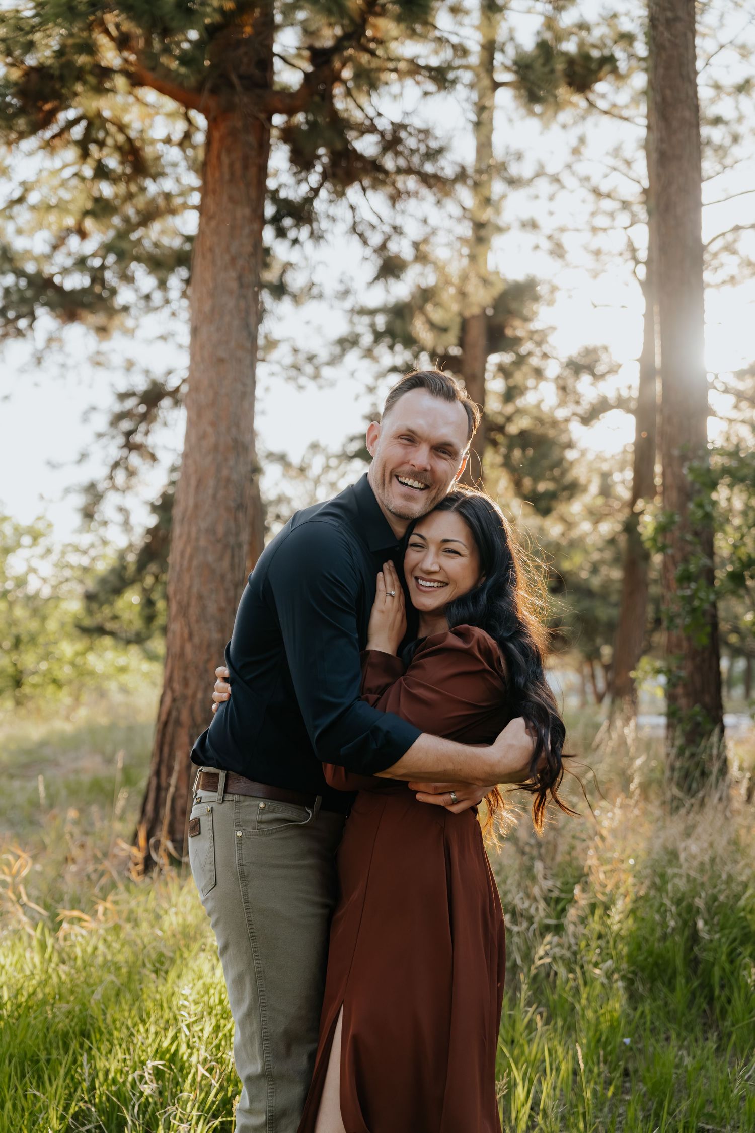 Couple embracing and smiling in a sunlit forest clearing, man in navy shirt hugging woman in rust dress.