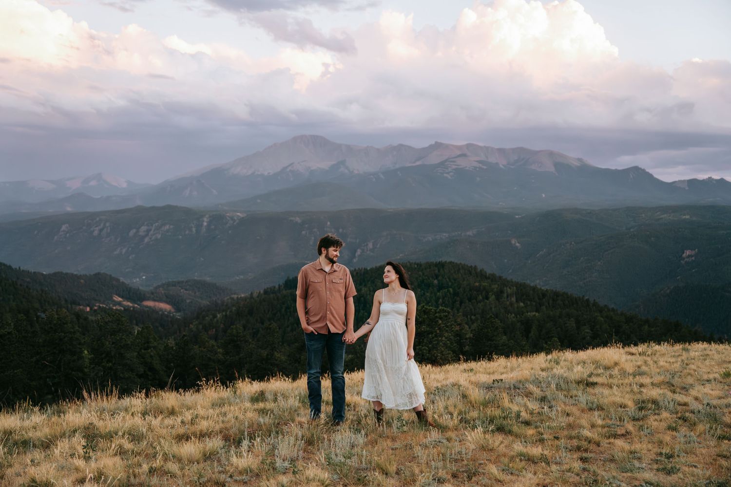 Couple holding hands on a mountain meadow at sunset with dramatic Rocky Mountain peaks and clouds in the background.