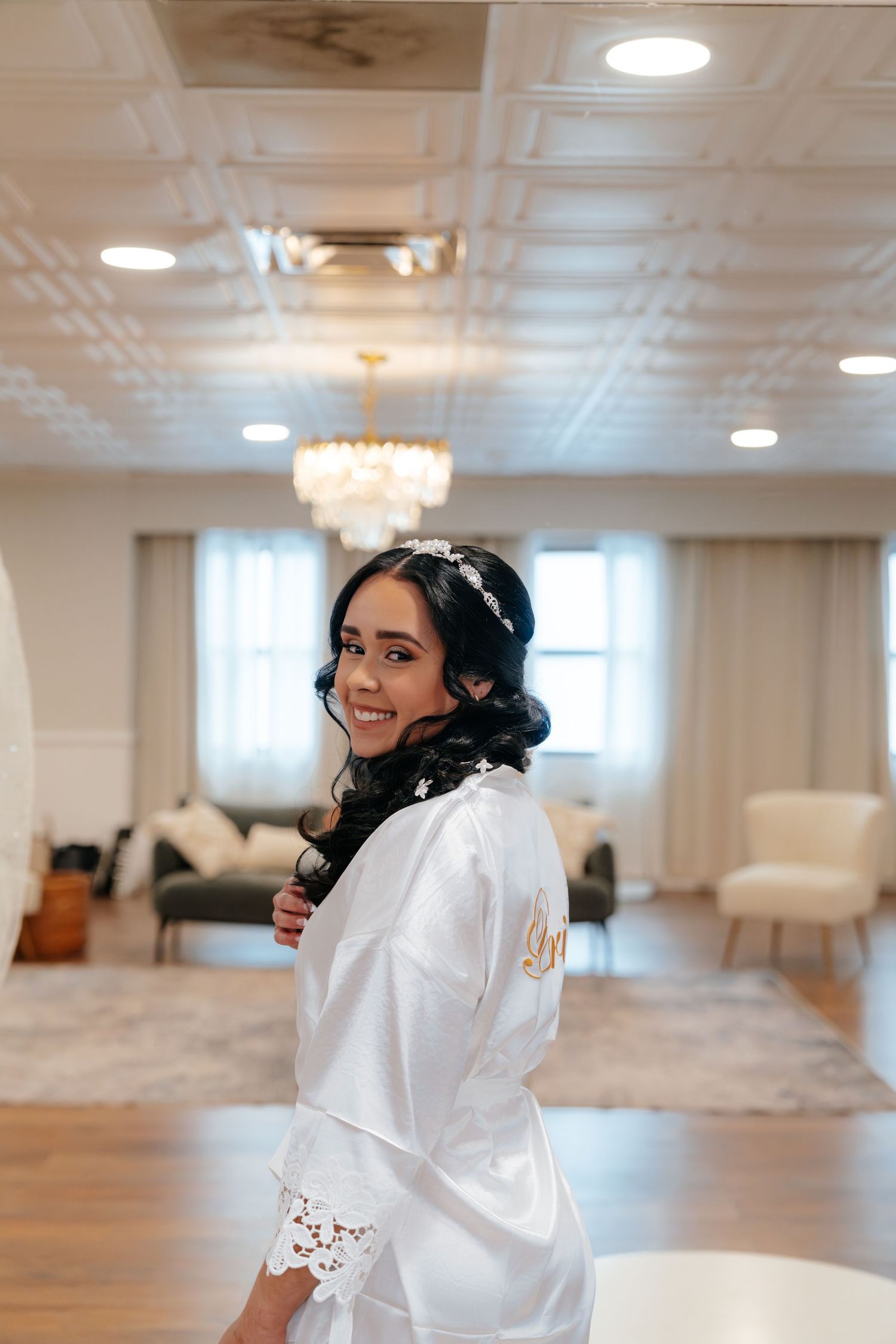 Smiling bride in white satin robe with lace trim and floral headpiece poses in elegant bridal suite.