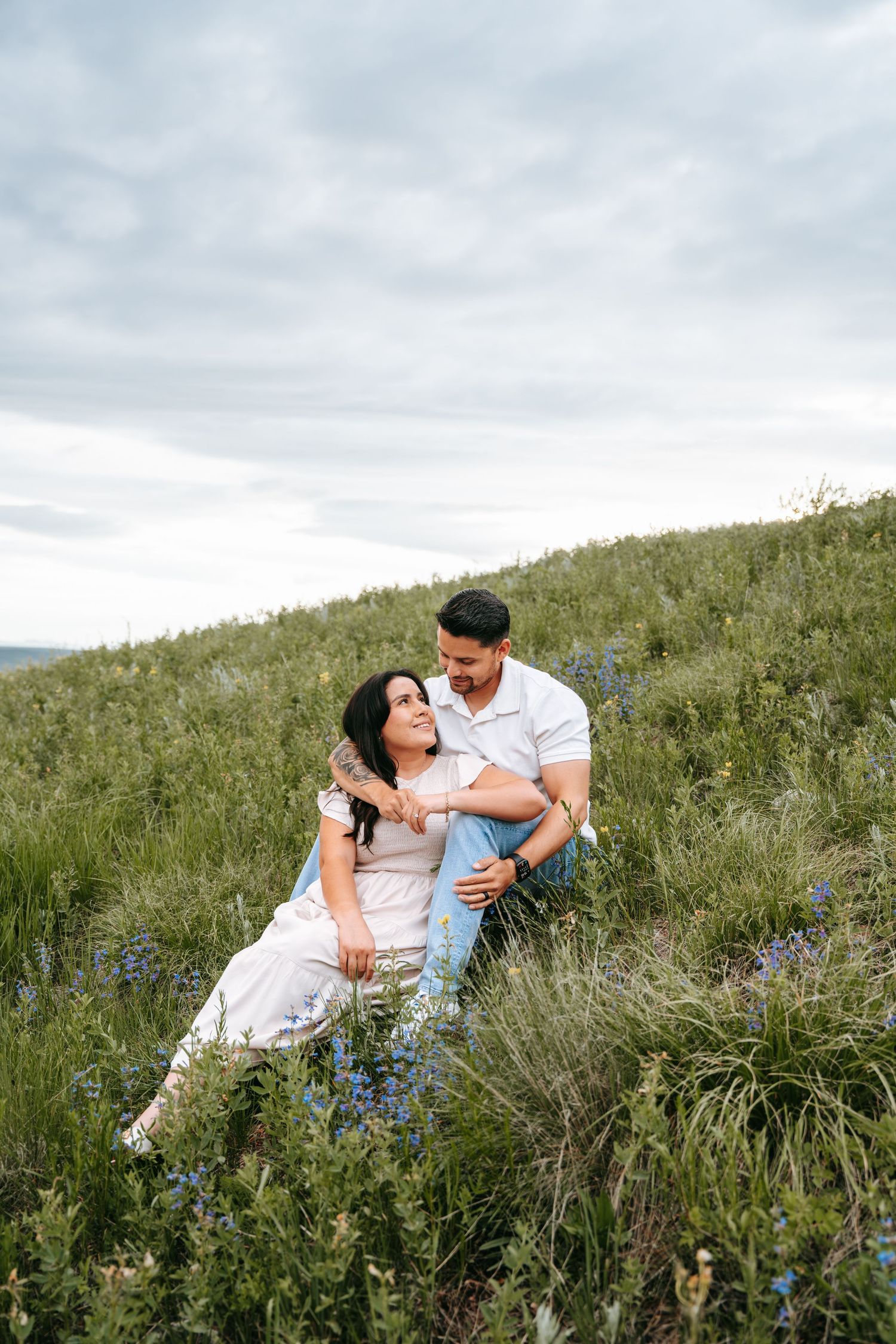 Couple relaxes together on a wildflower hillside, sharing a tender moment in green landscape.