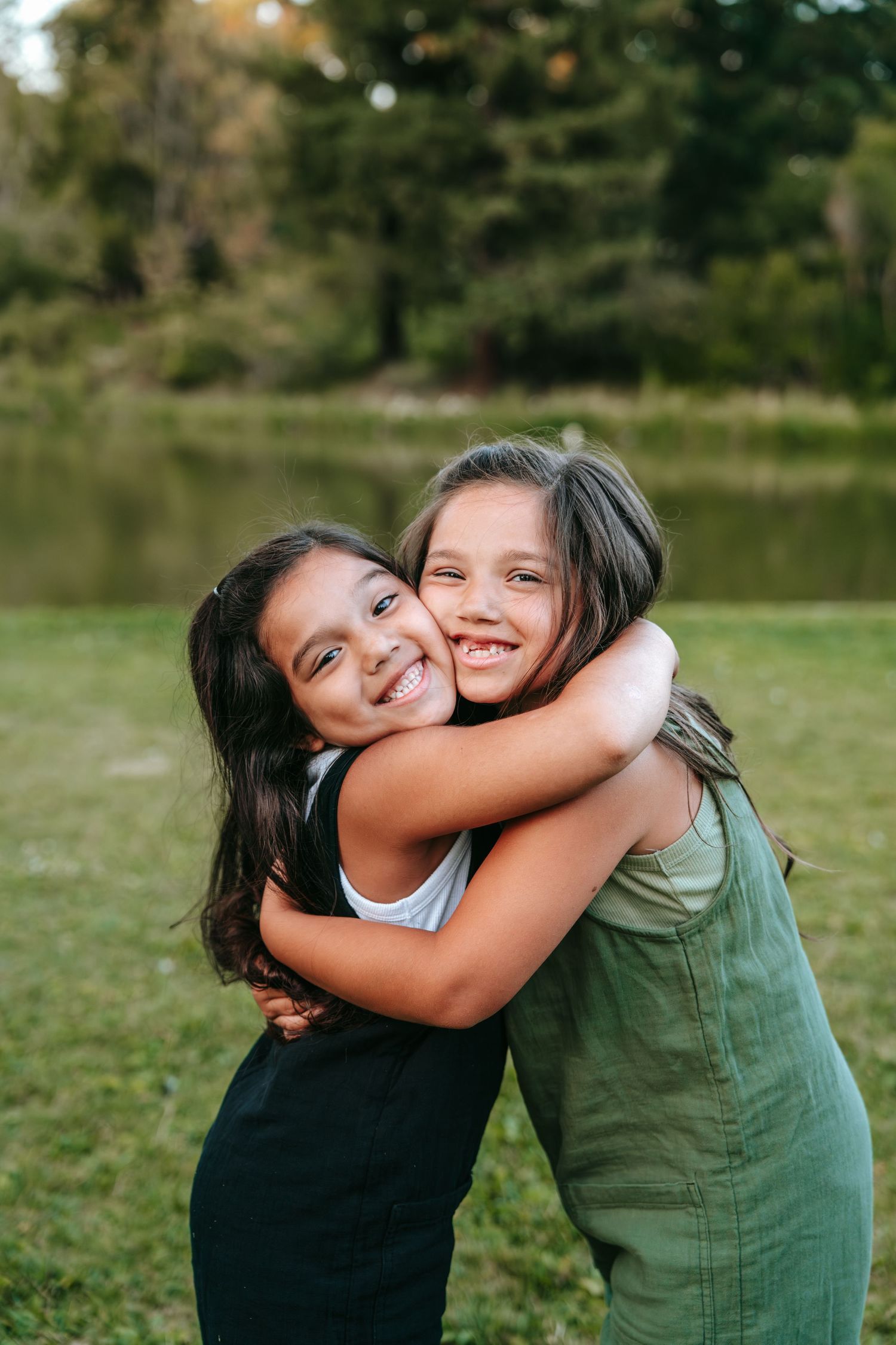Two young sisters smiling and hugging each other tightly in a lush green park by a pond.
