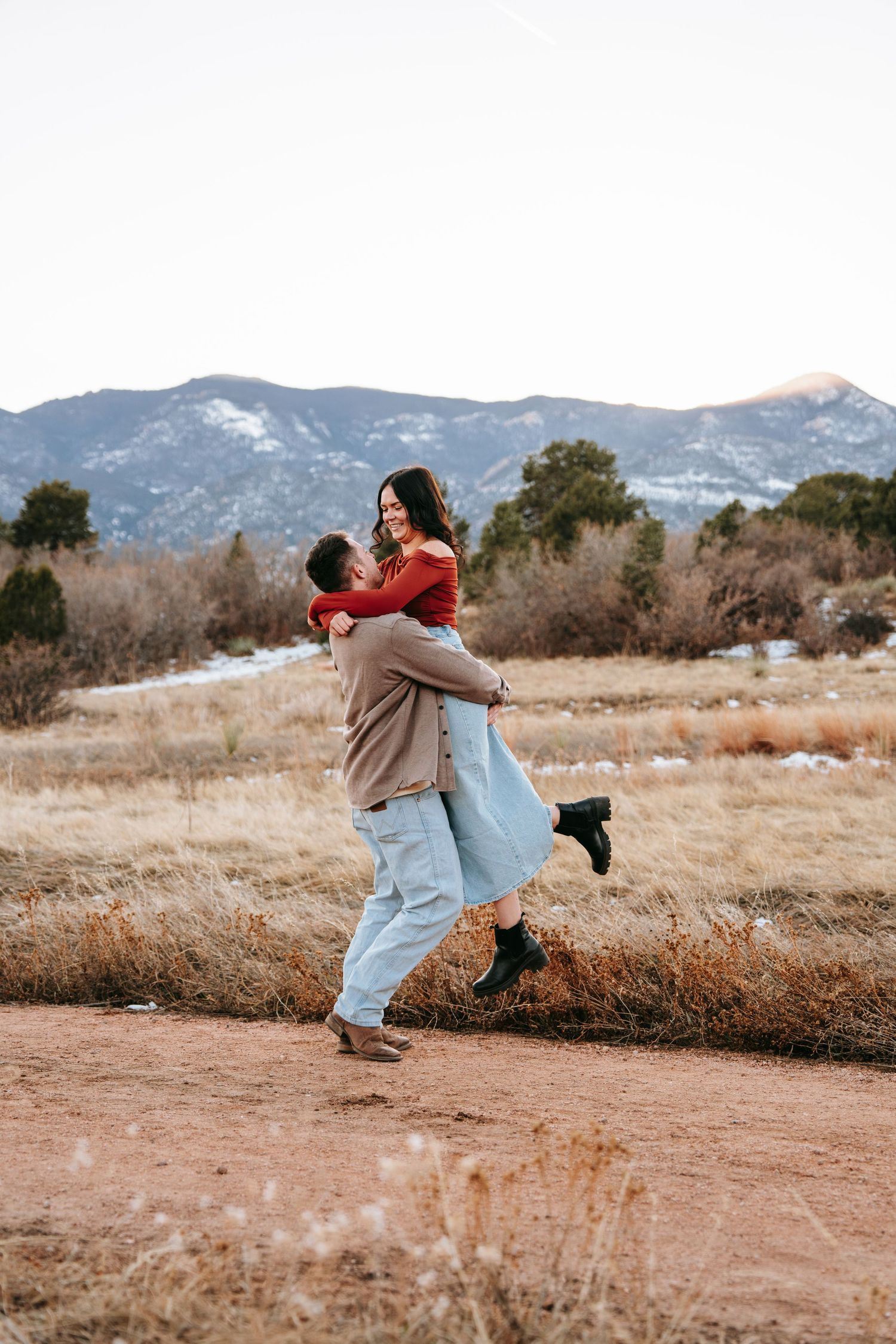 Man lifting and spinning woman in red top joyfully on a dirt path with mountains behind.