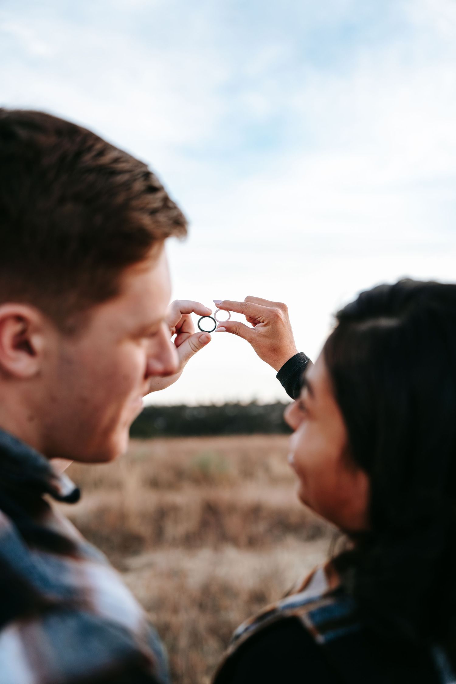 Couple facing each other holding up wedding rings between them in an outdoor field setting.