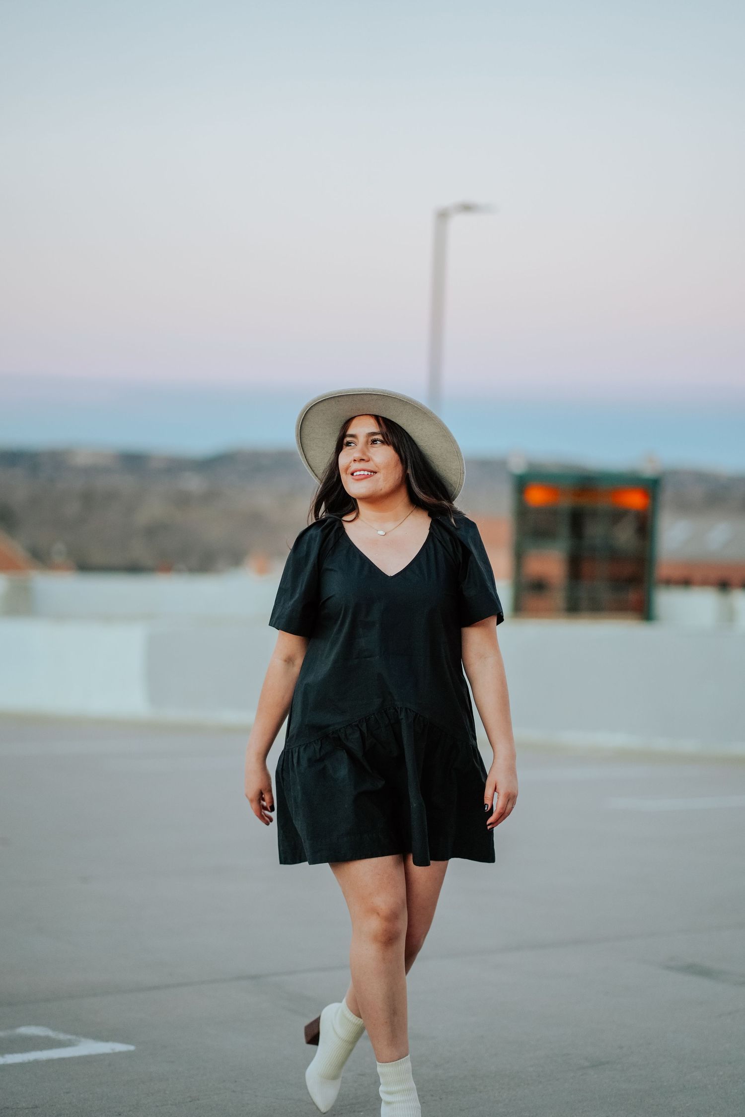 A young woman in a black dress and wide-brim hat walks on a rooftop at dusk.
