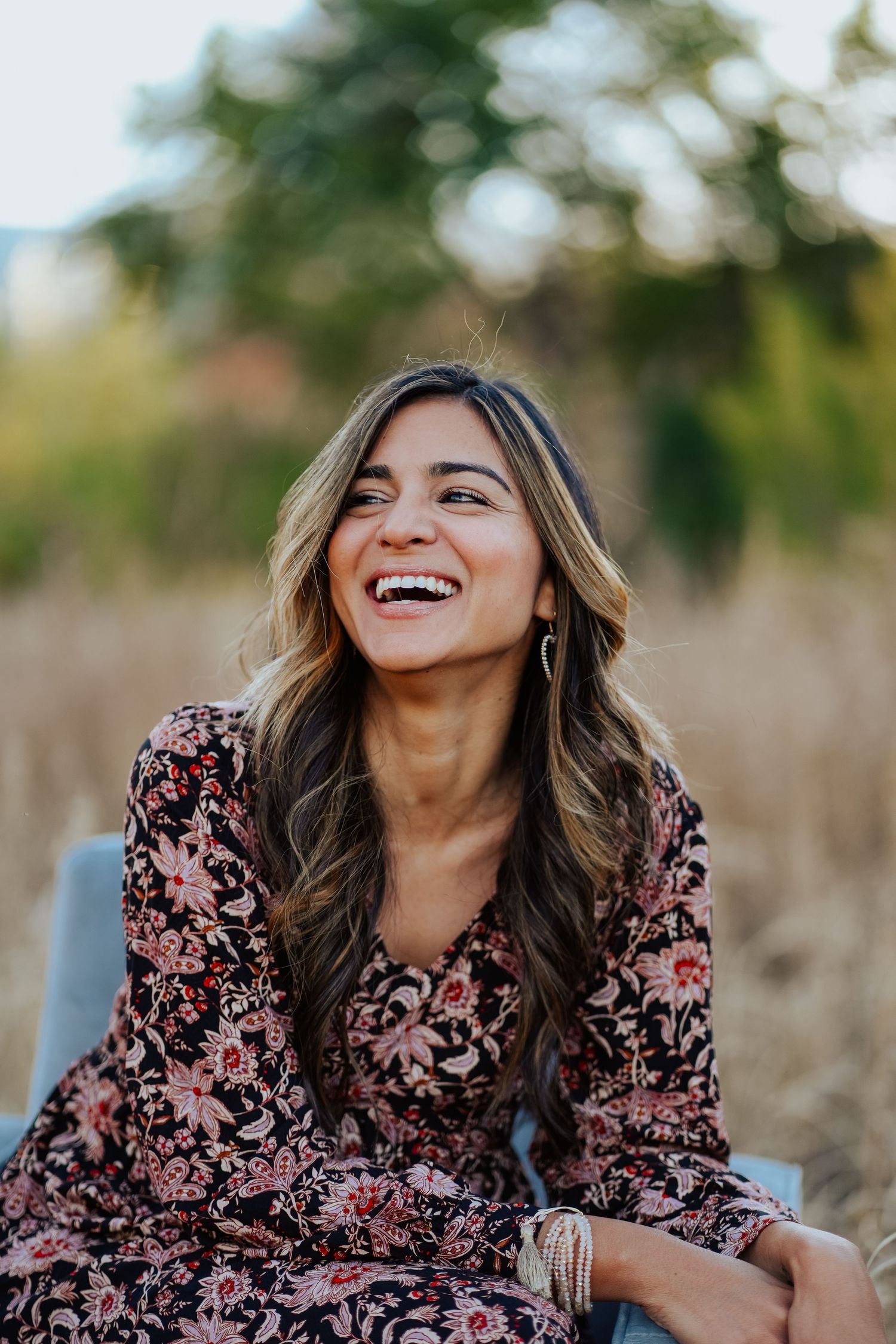 Young woman with long wavy hair in floral dress laughing joyfully outdoors in golden field.