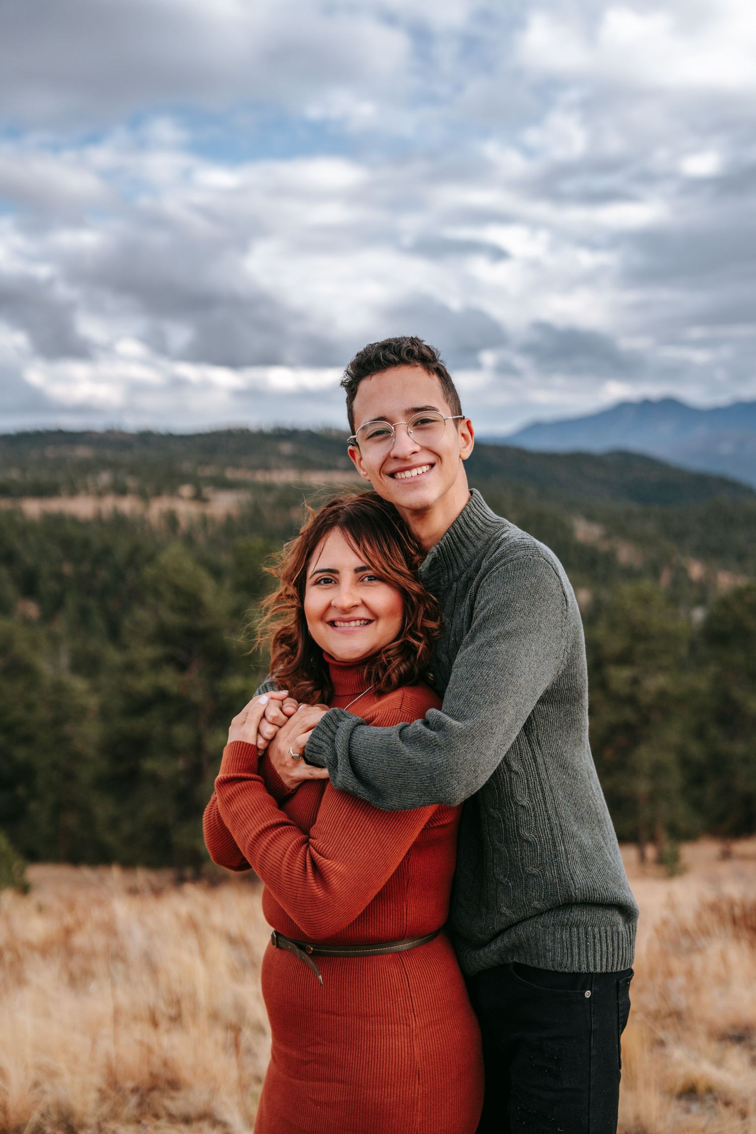 Young man hugging woman from behind while smiling in a scenic mountain landscape with cloudy skies.