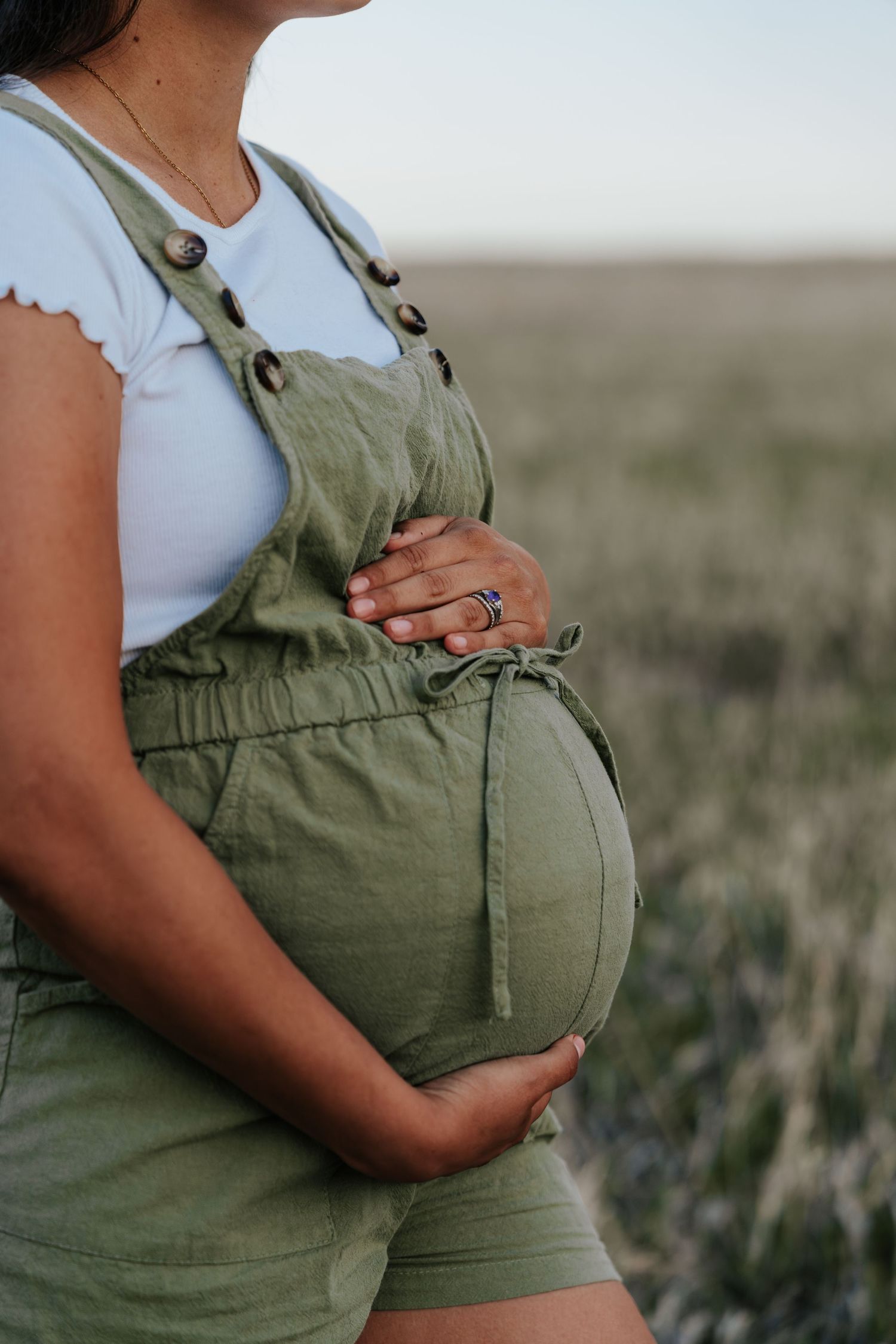 Close-up of pregnant woman in green overalls cradling baby bump in a field.
