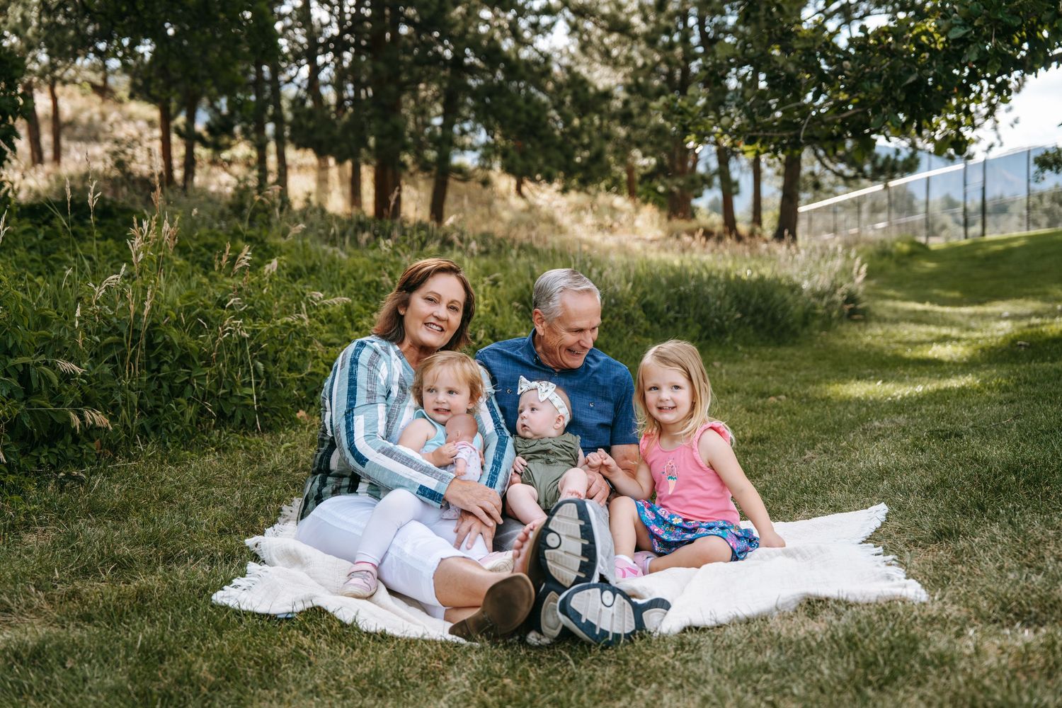 Family of grandparents and three young grandchildren smiling together on a blanket in a lush green park.