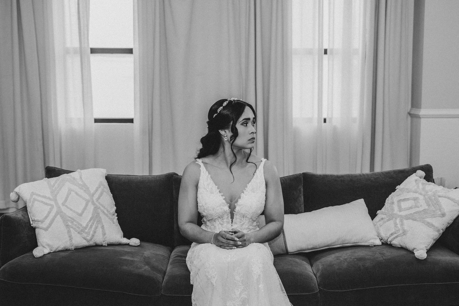 Black and white photo of a bride in a lace wedding dress sitting on a sofa, looking contemplative before her ceremony.