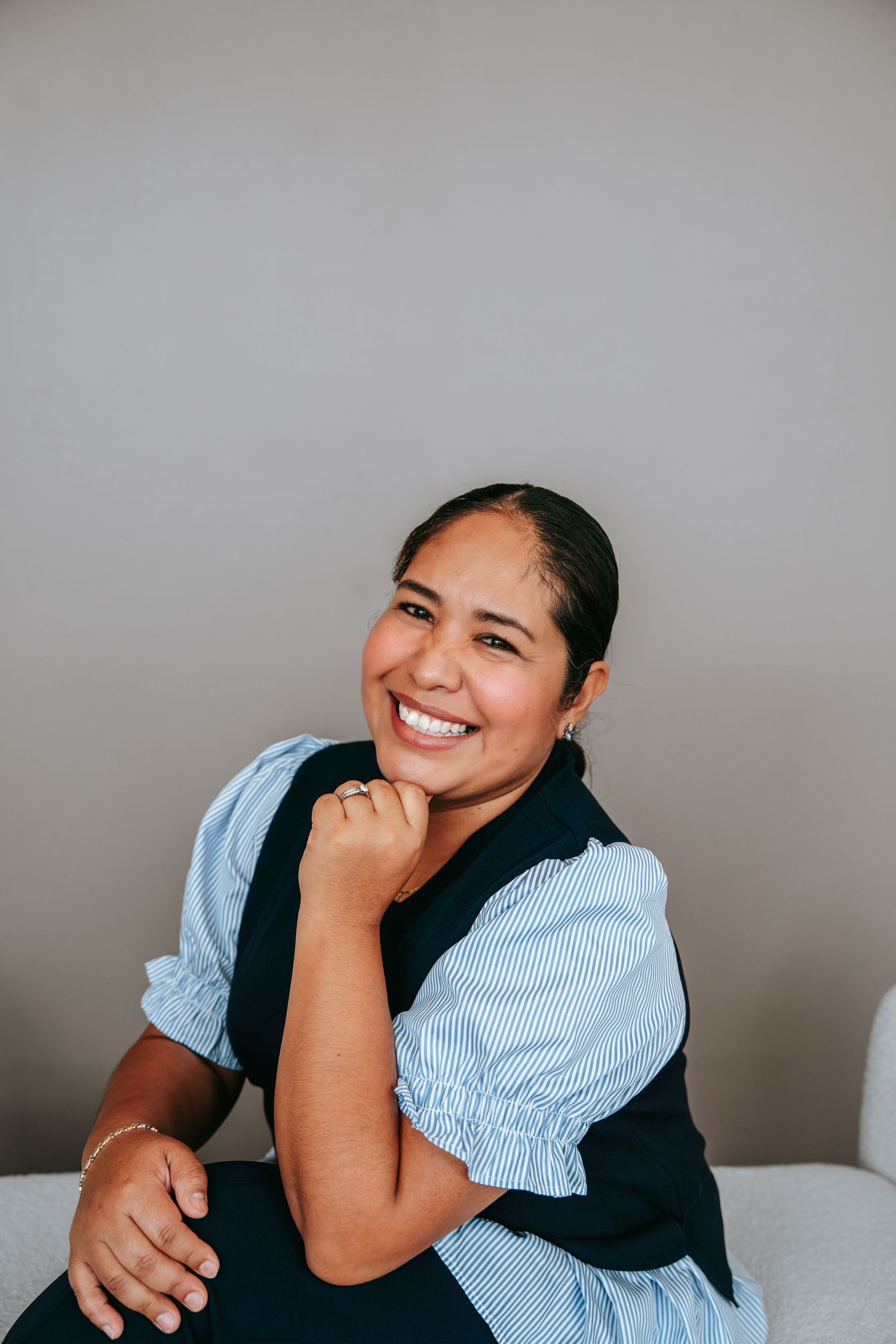 Smiling woman in a blue striped shirt and black vest sitting with hand on chin against gray background.
