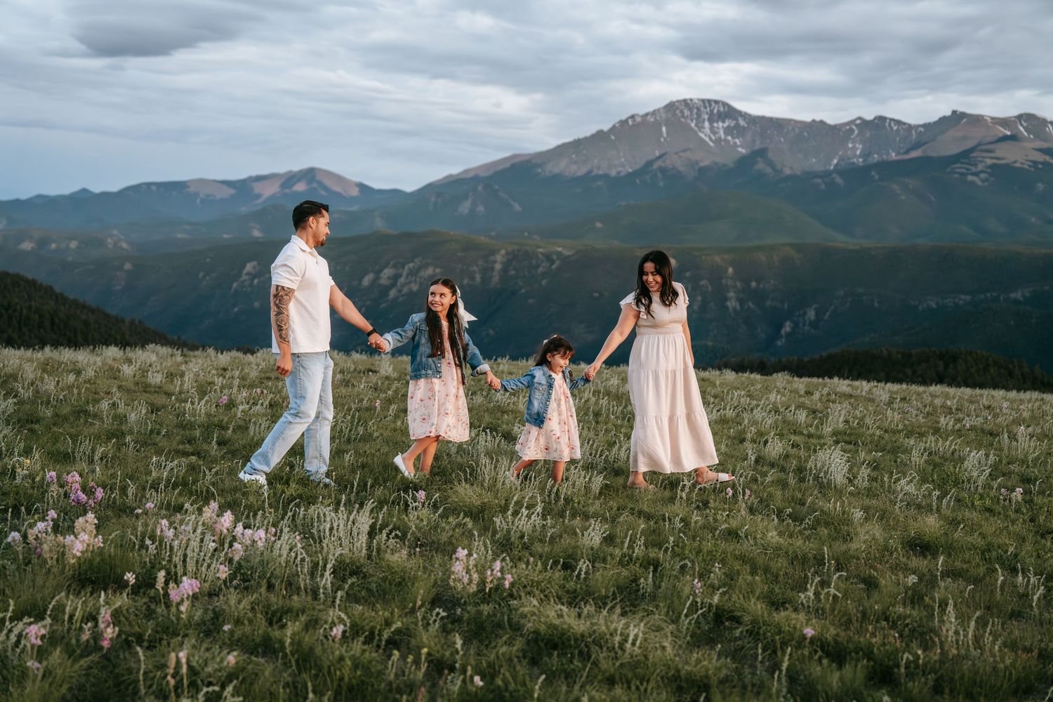 Happy family of four holding hands walking through a wildflower meadow with mountain backdrop.