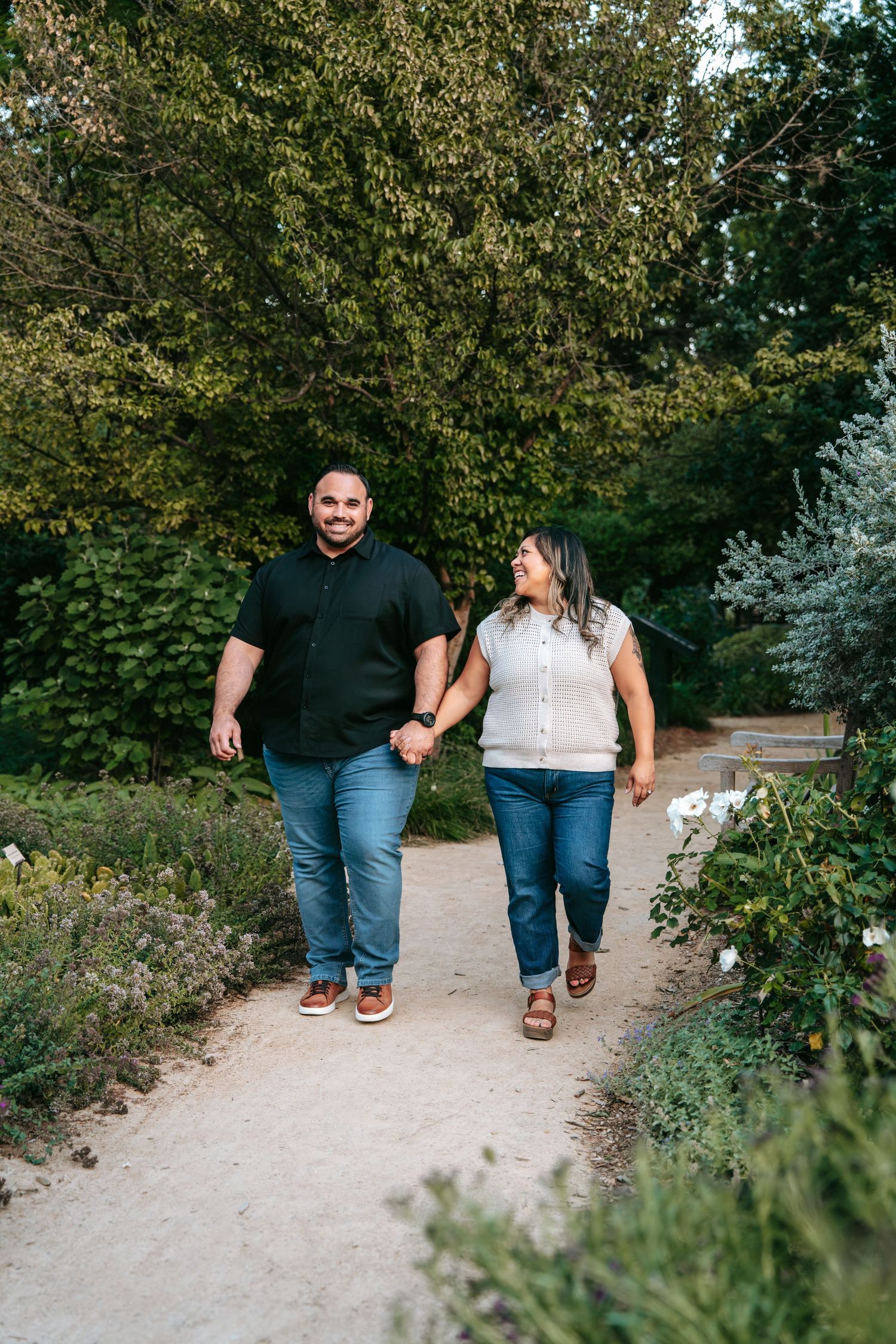 A smiling couple holds hands while walking along a garden path surrounded by lush green trees and plants.