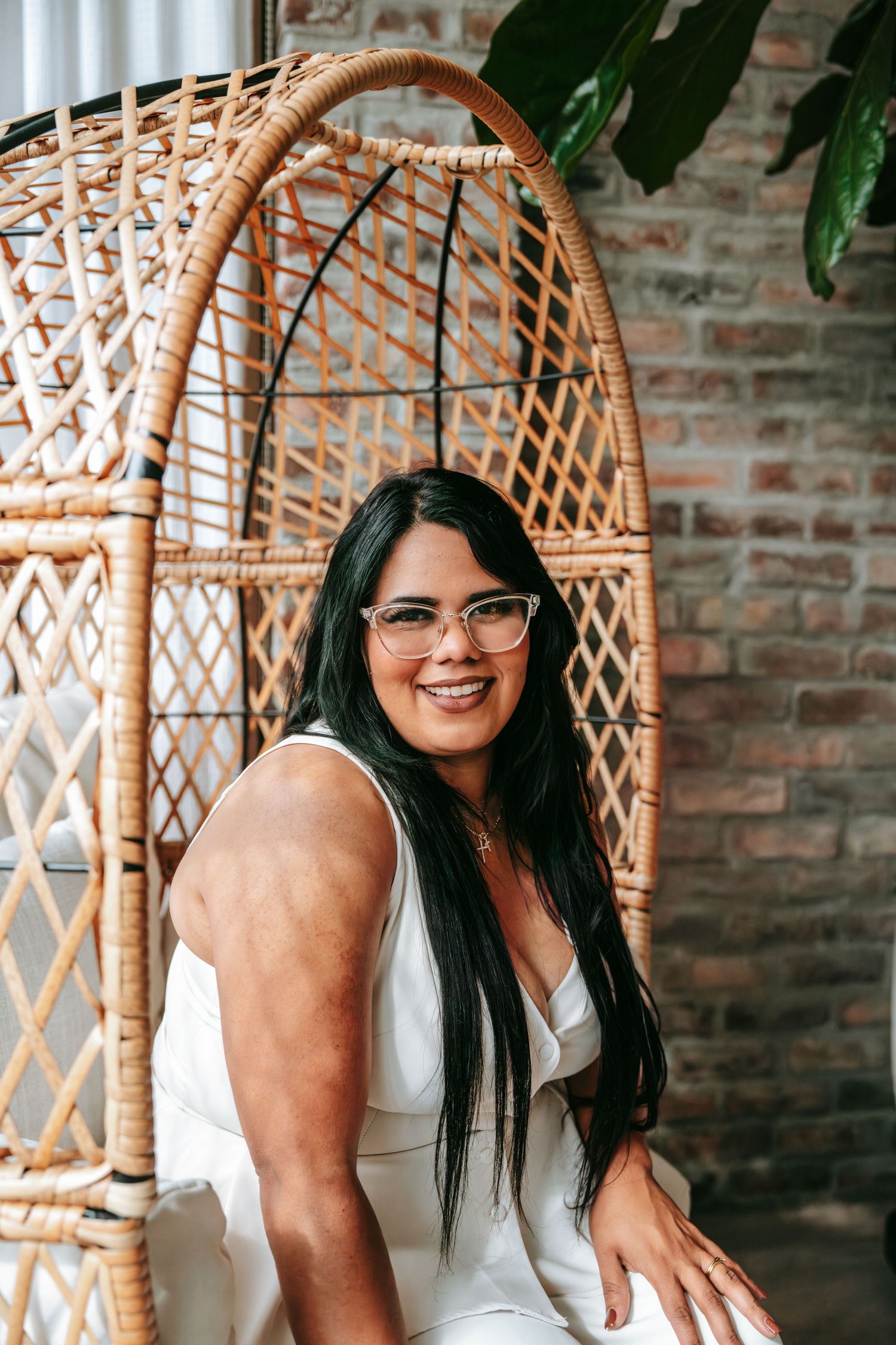A smiling woman with long dark hair and glasses sits in a large wicker egg chair against a brick wall.