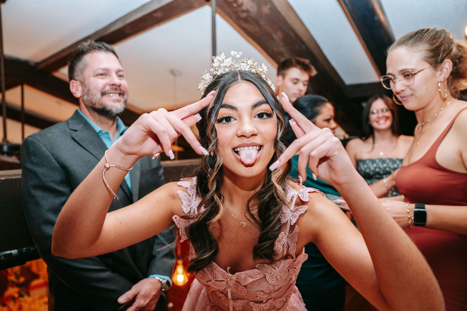 Quinceañera girl in pink gown and crown making fun poses with guests at celebration party.