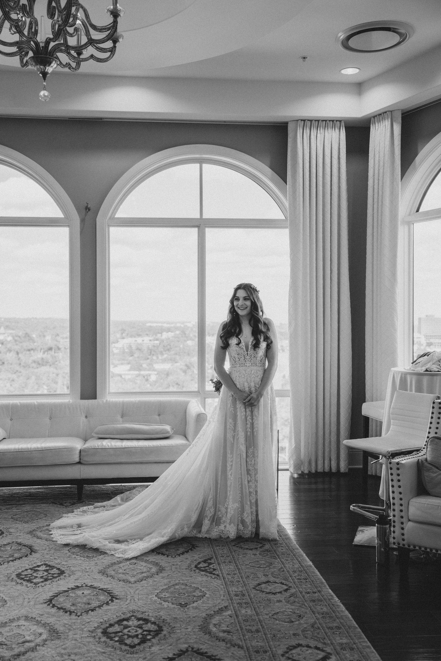 Elegant black and white portrait of bride in lace gown standing by arched windows in a luxurious suite.