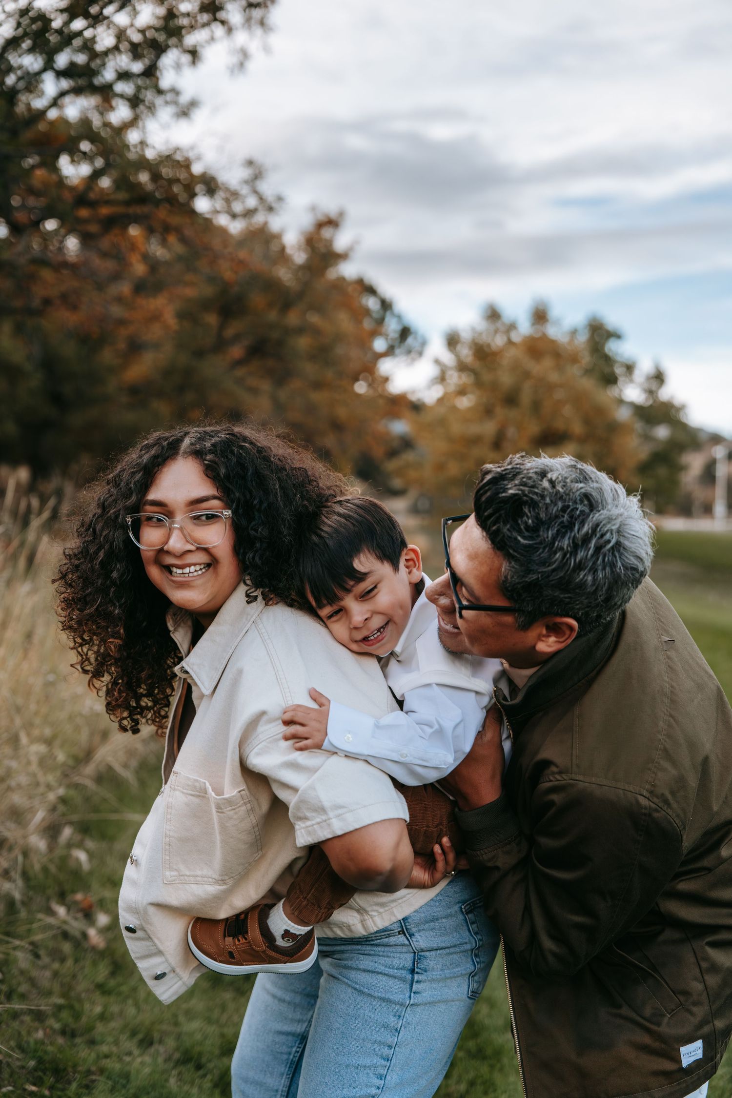 Laughing family of three playing together in a grassy field with colorful autumn trees behind them.