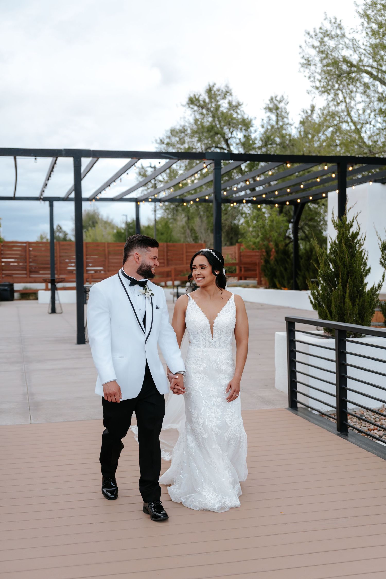 Bride and groom holding hands on outdoor deck, groom in white tuxedo jacket, bride in lace fitted wedding gown.
