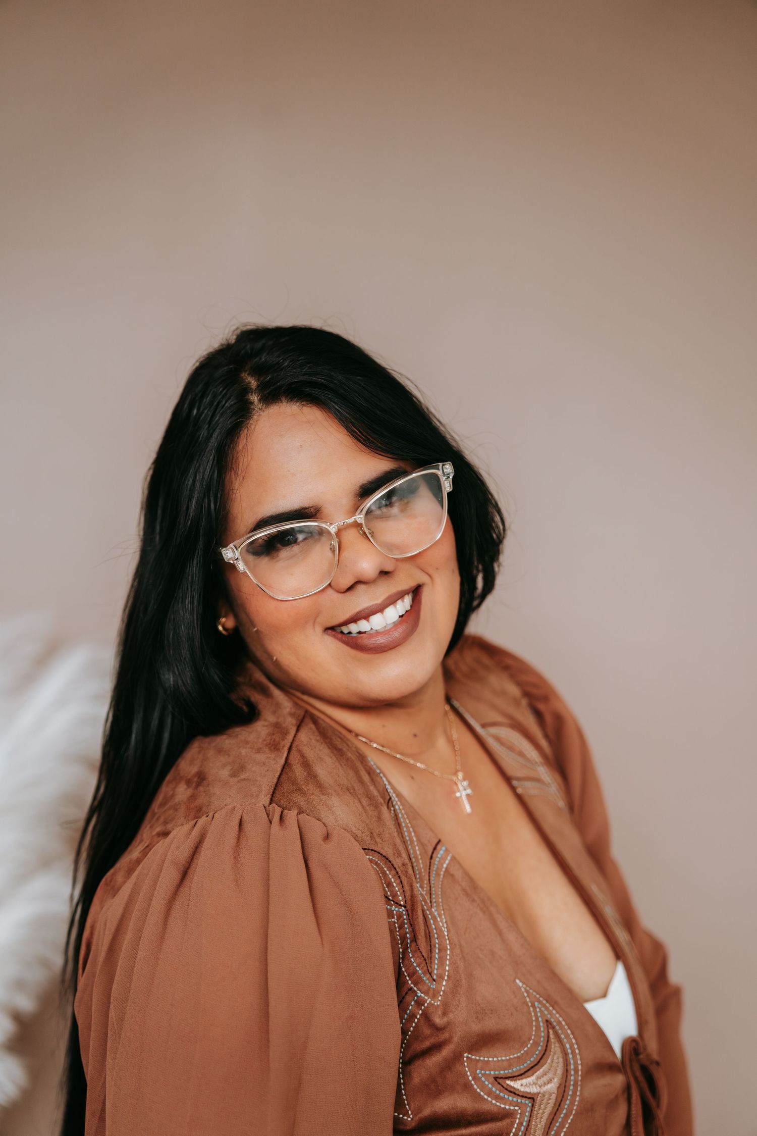 Smiling woman with glasses wearing a brown blouse and cross necklace posing against a beige background.
