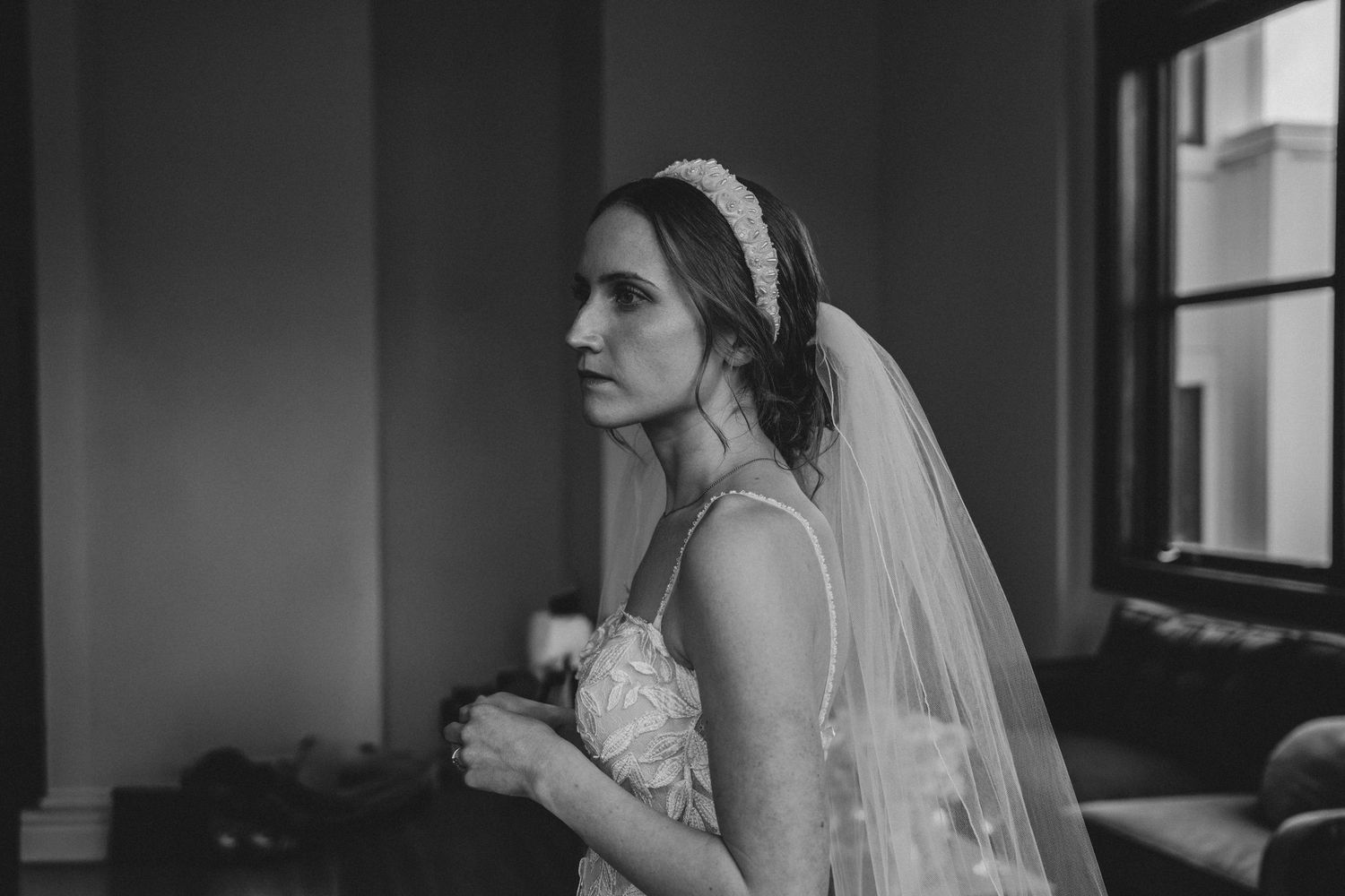 Black and white portrait of a bride in a veil and lace dress gazing out a window on her wedding day.