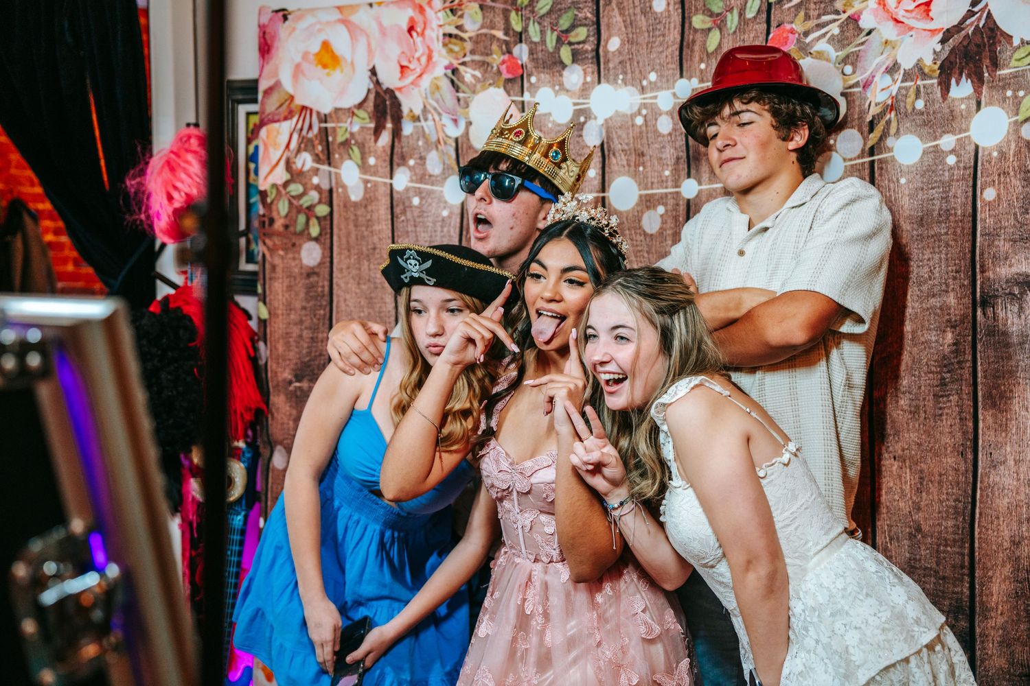 Group of teens posing with fun props at quinceañera photo booth with floral wood backdrop.