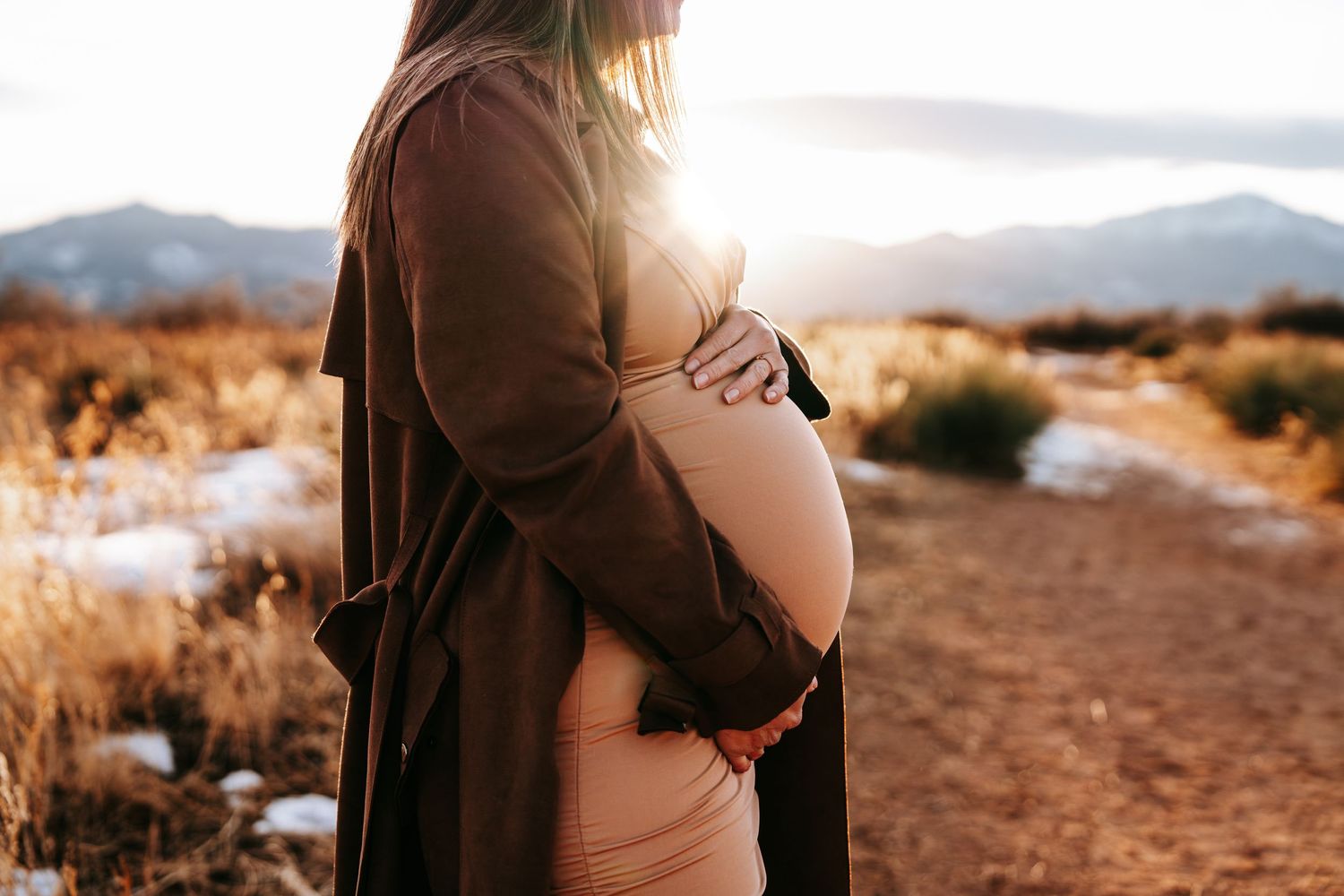 Close-up of pregnant woman in pink outfit and brown cardigan cradling bare baby bump at golden hour.