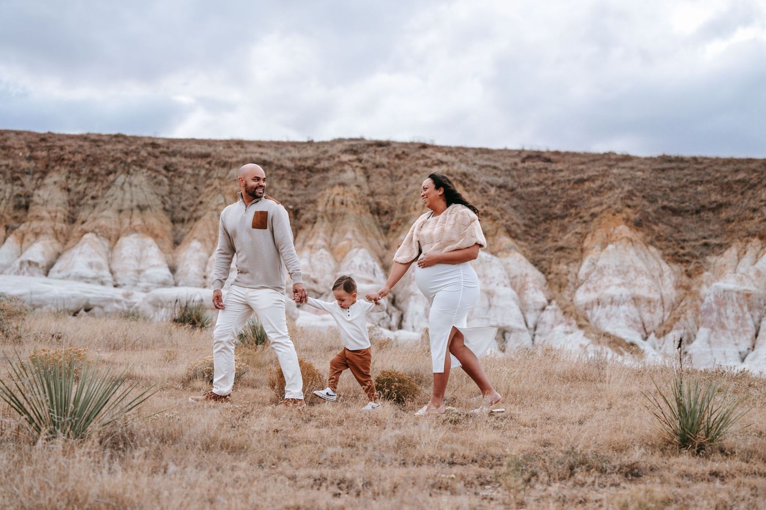 Family of three holding hands and walking through dry golden grass with desert hoodoos behind.