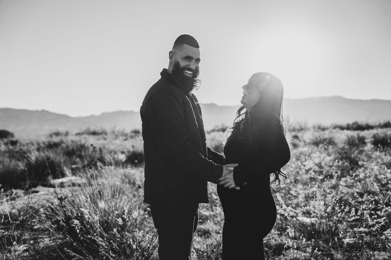 Black and white maternity photo of couple holding hands in sunlit field with mountain landscape.