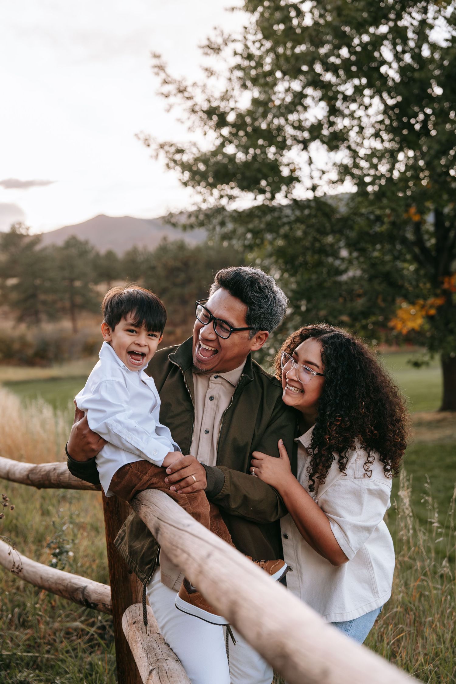 Smiling family of three enjoying time together leaning on a wooden fence in a scenic park.