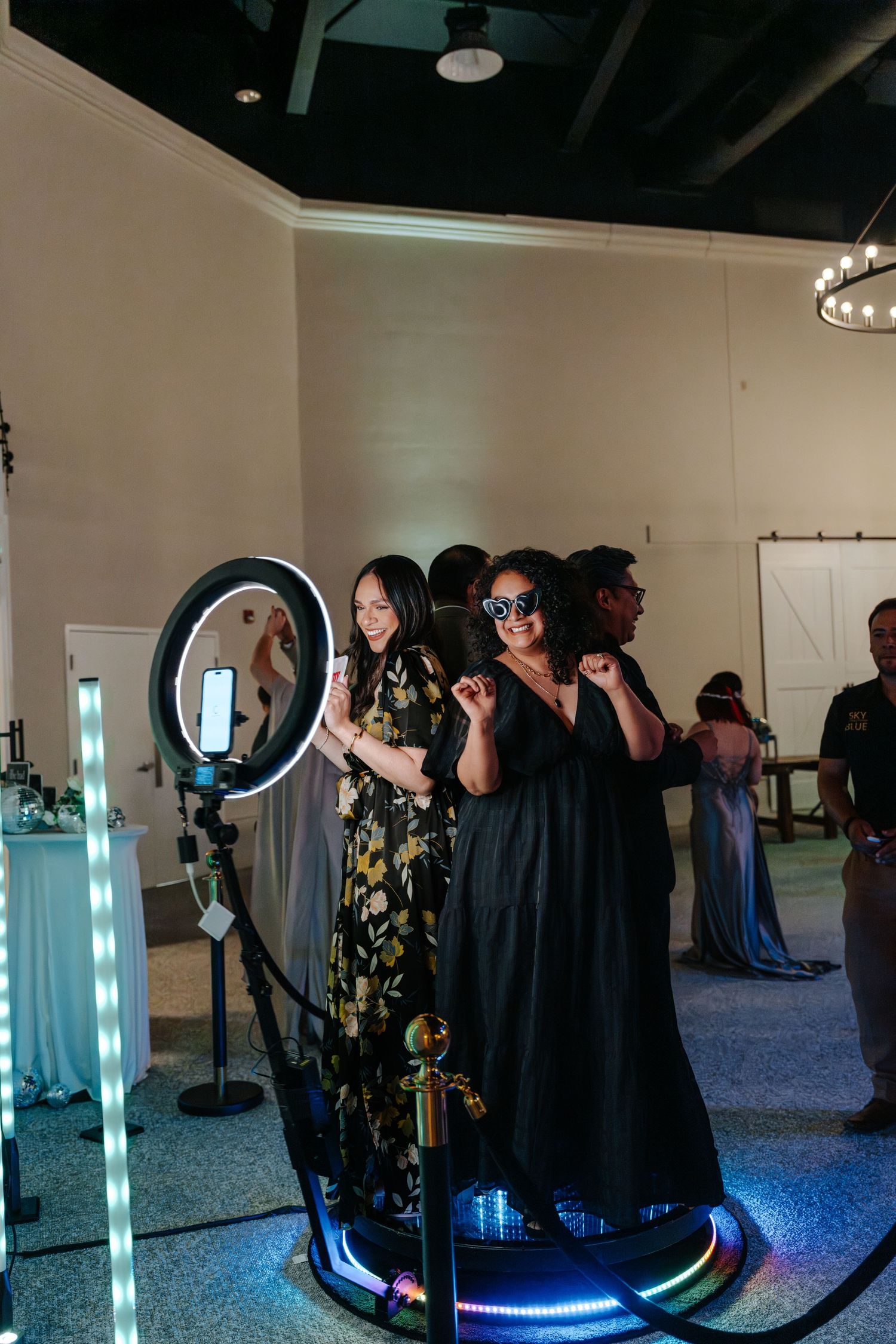 Two women posing at a ring light photo booth setup at an elegant indoor wedding reception venue.