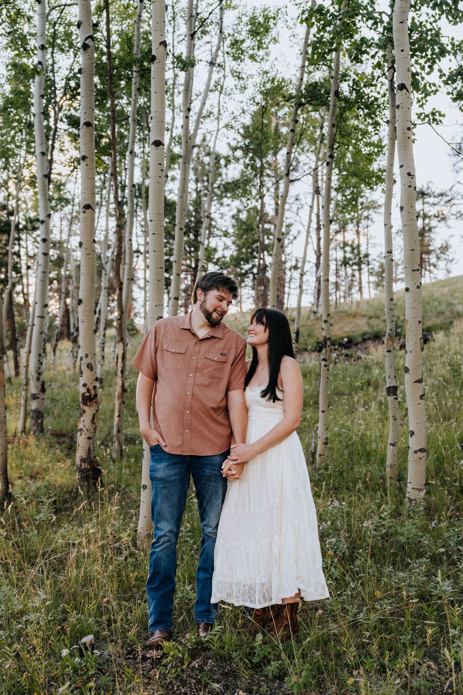 Couple holding hands and smiling among white aspen trees in a lush green forest clearing.