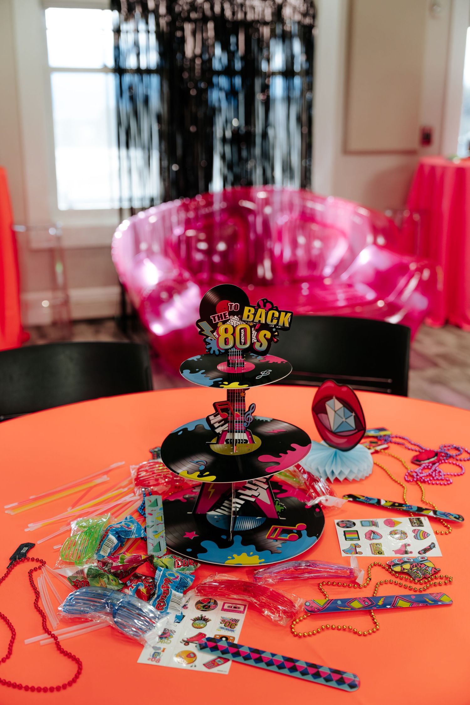 80s themed party table with colorful favors, tiered display, beads, and stickers on an orange tablecloth.