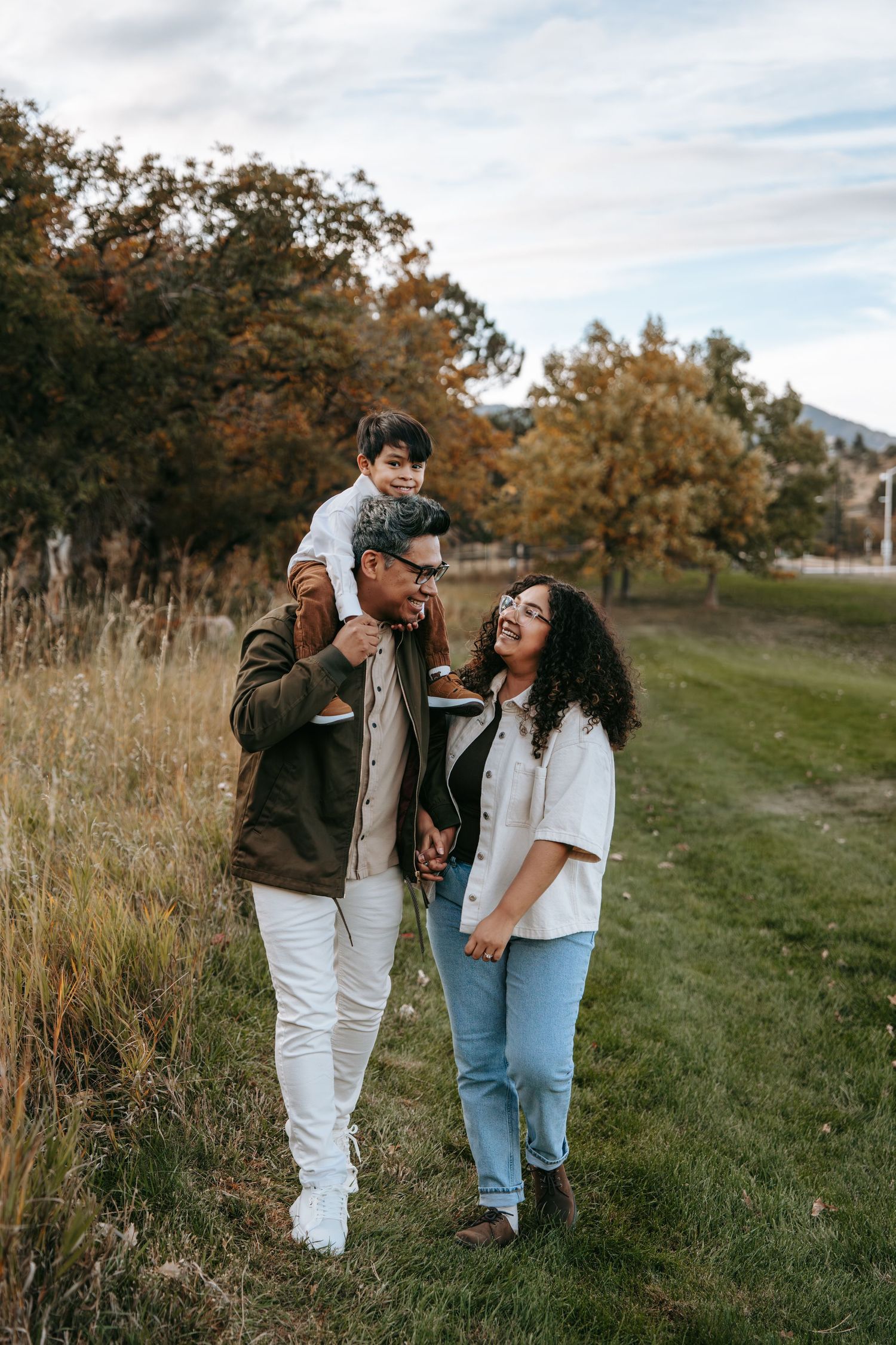 Happy family walking through autumn meadow with child riding on fathers shoulders in fall landscape.