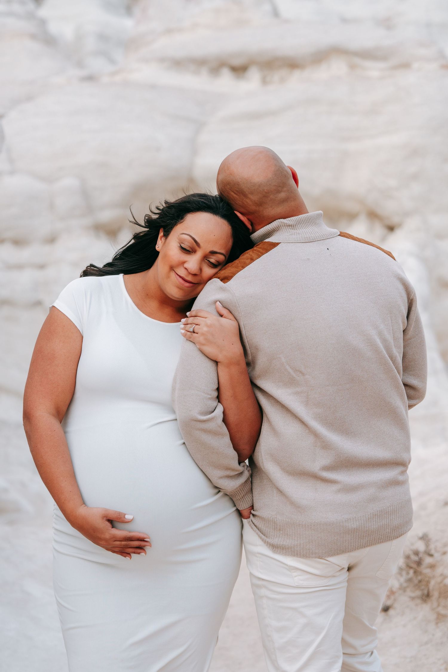 Pregnant woman in white dress leaning on partner near white rock formations during maternity shoot.