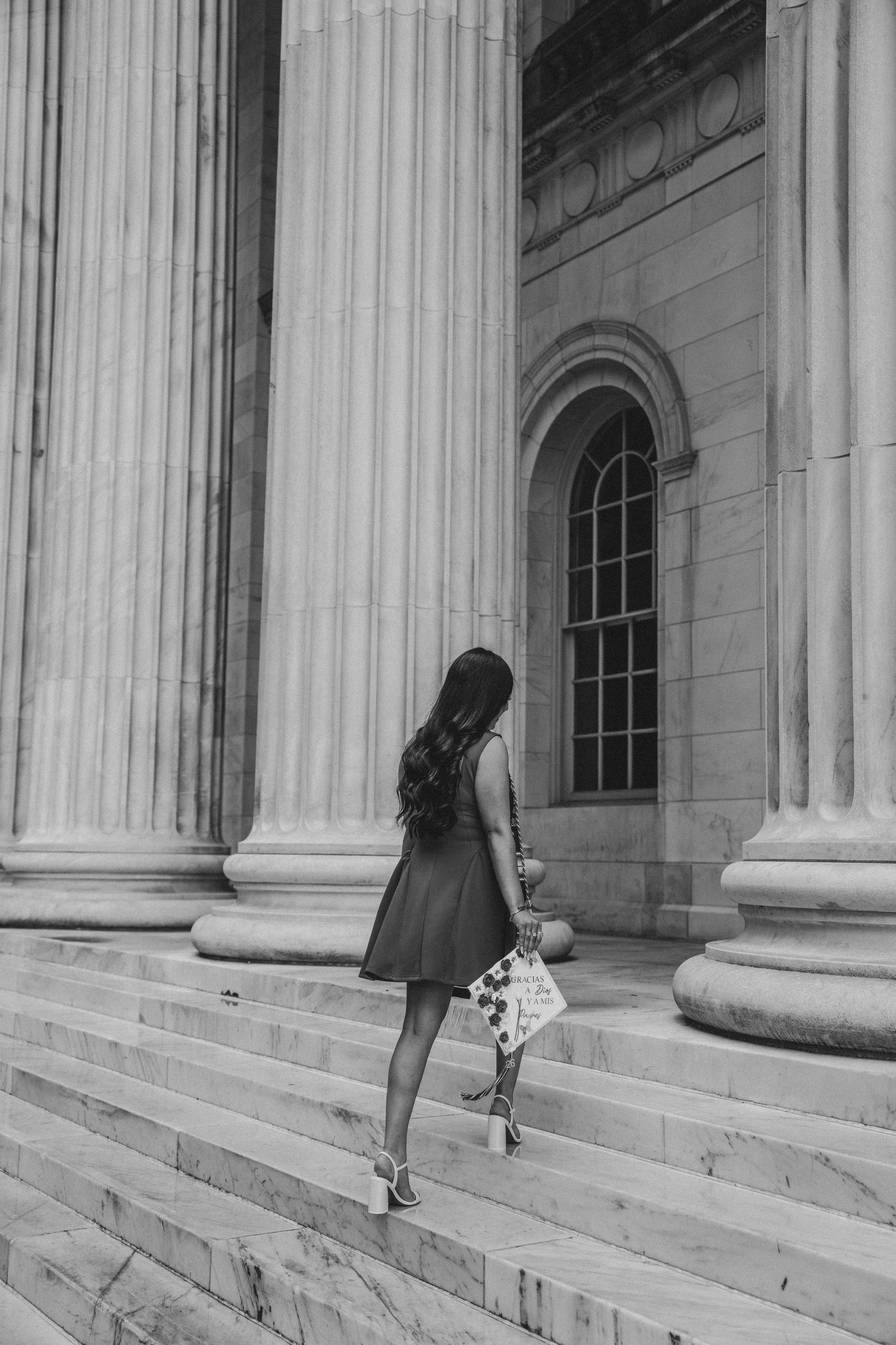Black and white rear view of a young woman in red dress carrying decorated graduation cap on stone steps.