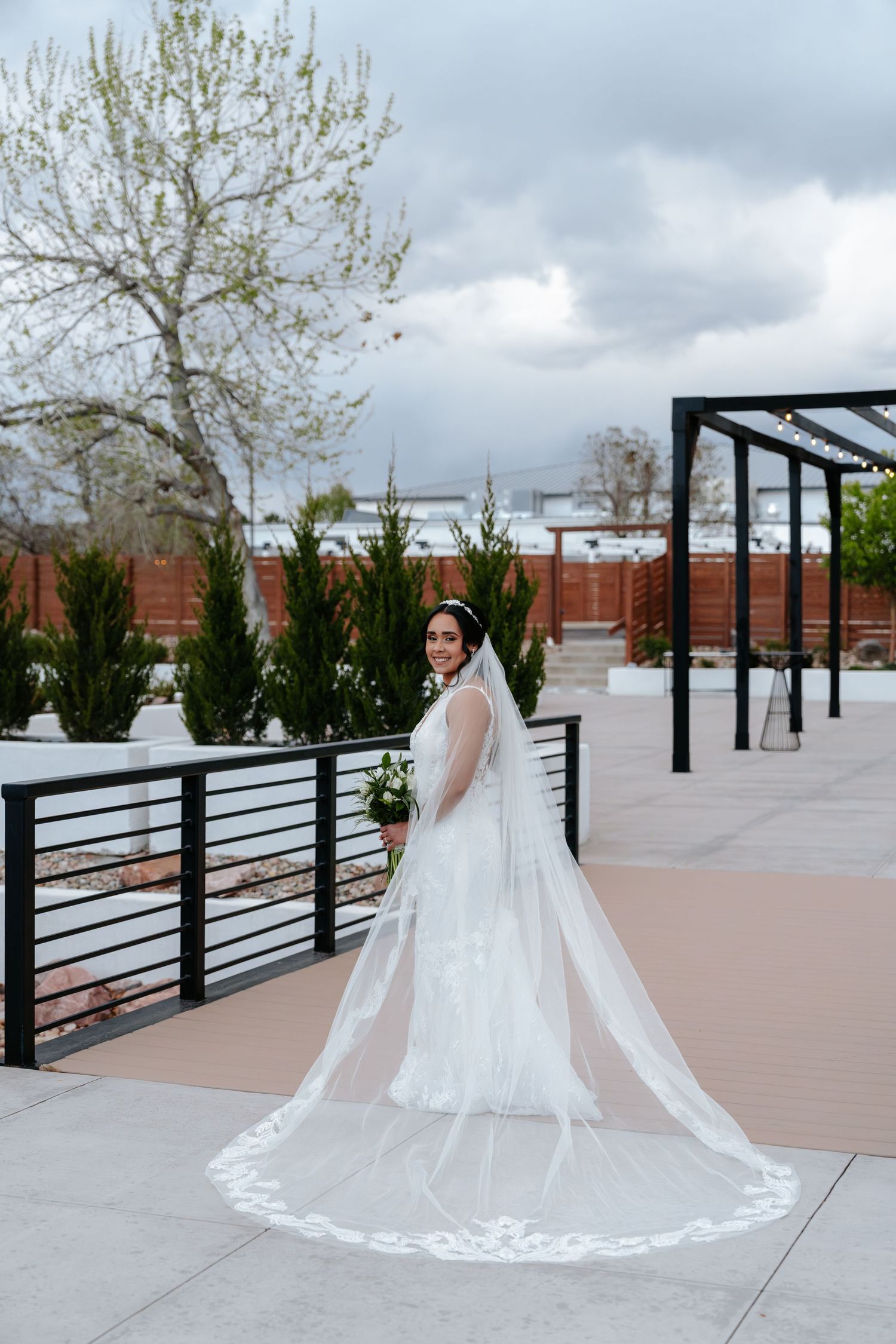Bride in flowing white gown with long lace-trimmed cathedral veil poses on outdoor wedding venue patio.