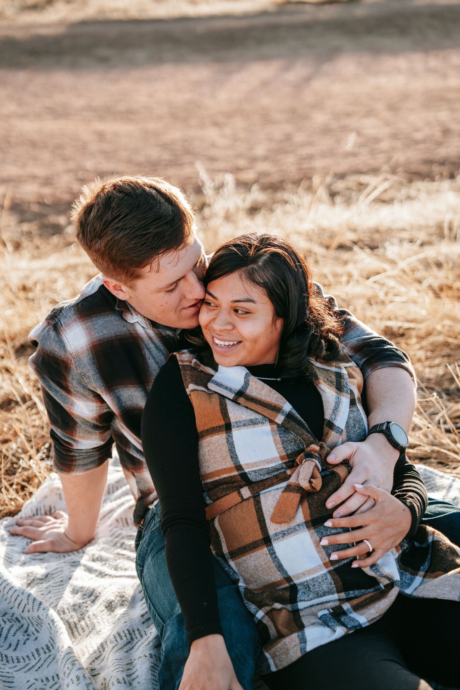 Couple sitting on a blanket in a field, man kissing woman's cheek as she smiles during maternity shoot.