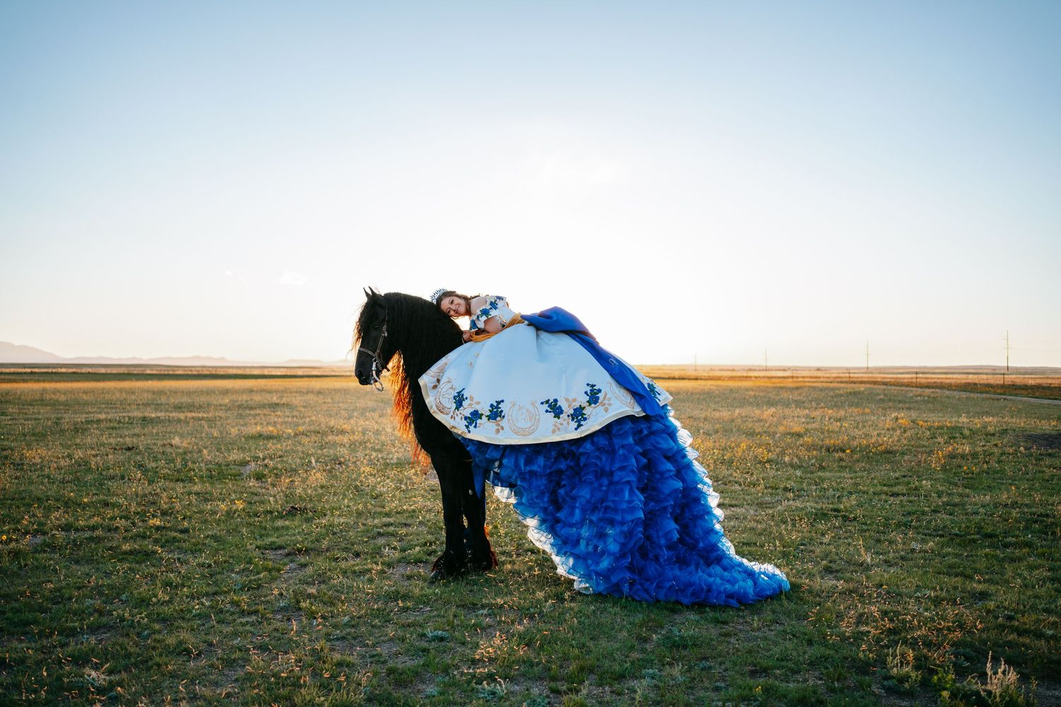 Young woman in blue quinceañera gown poses with black horse in open field at golden hour.