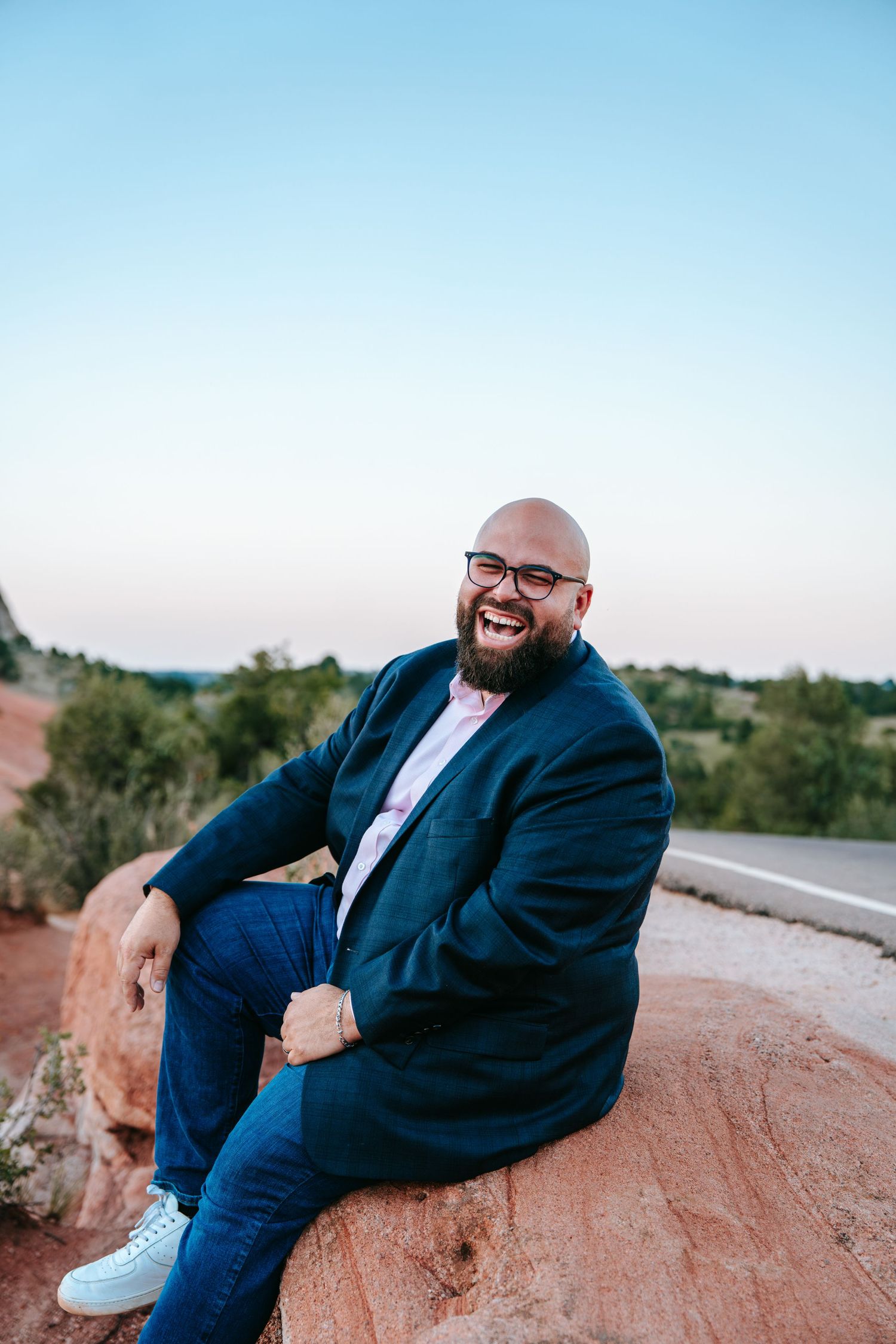 Bald bearded man in navy blazer laughing while sitting on red rocks with scenic landscape background.