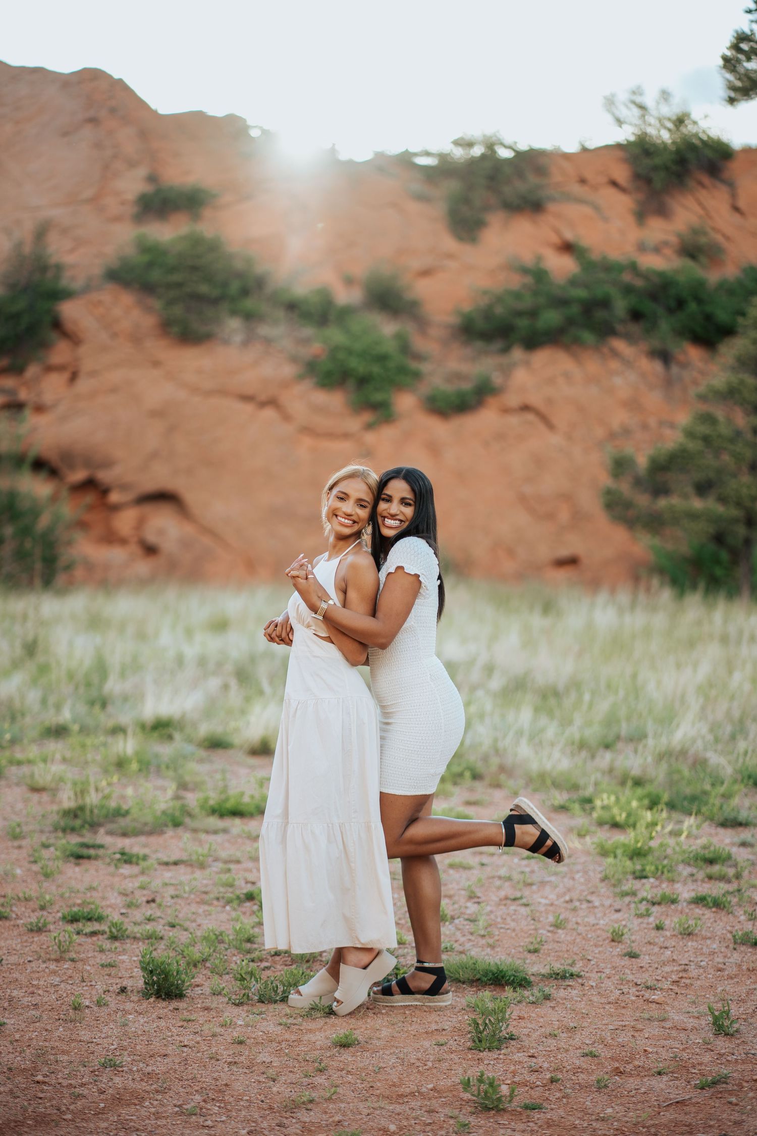 Two women in white dresses embrace and smile at red rock park in Colorado.