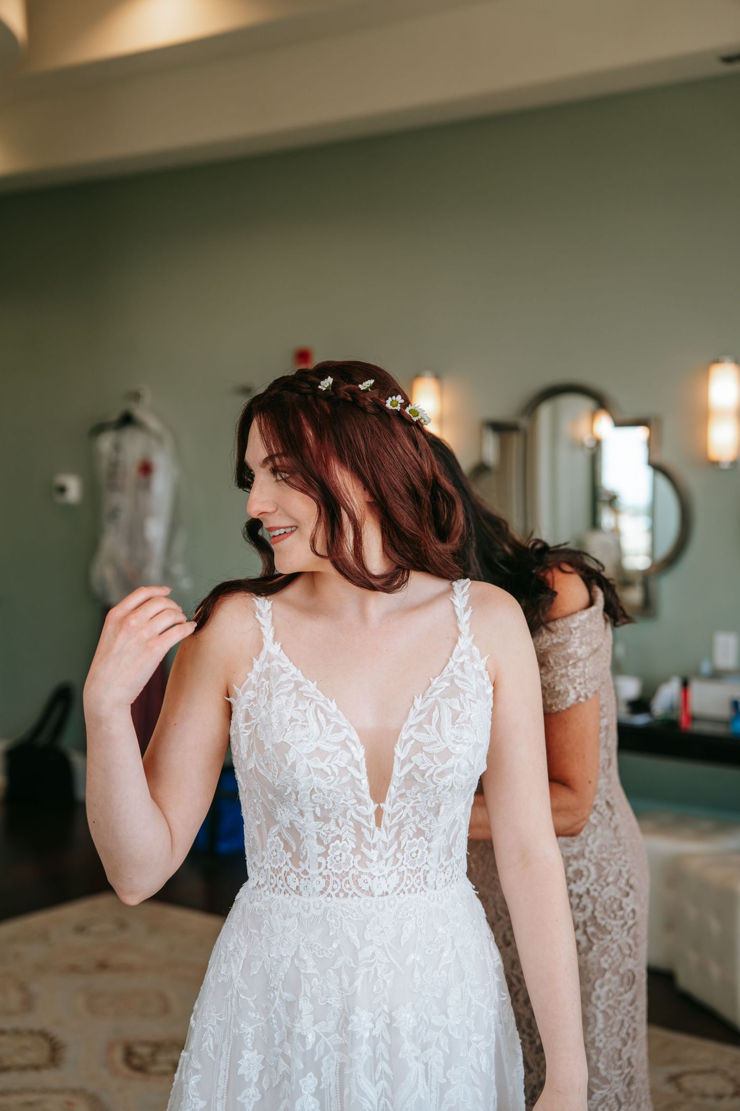 Smiling bride with auburn hair and floral hair pins adjusts her lace V-neck wedding dress in a green dressing room.