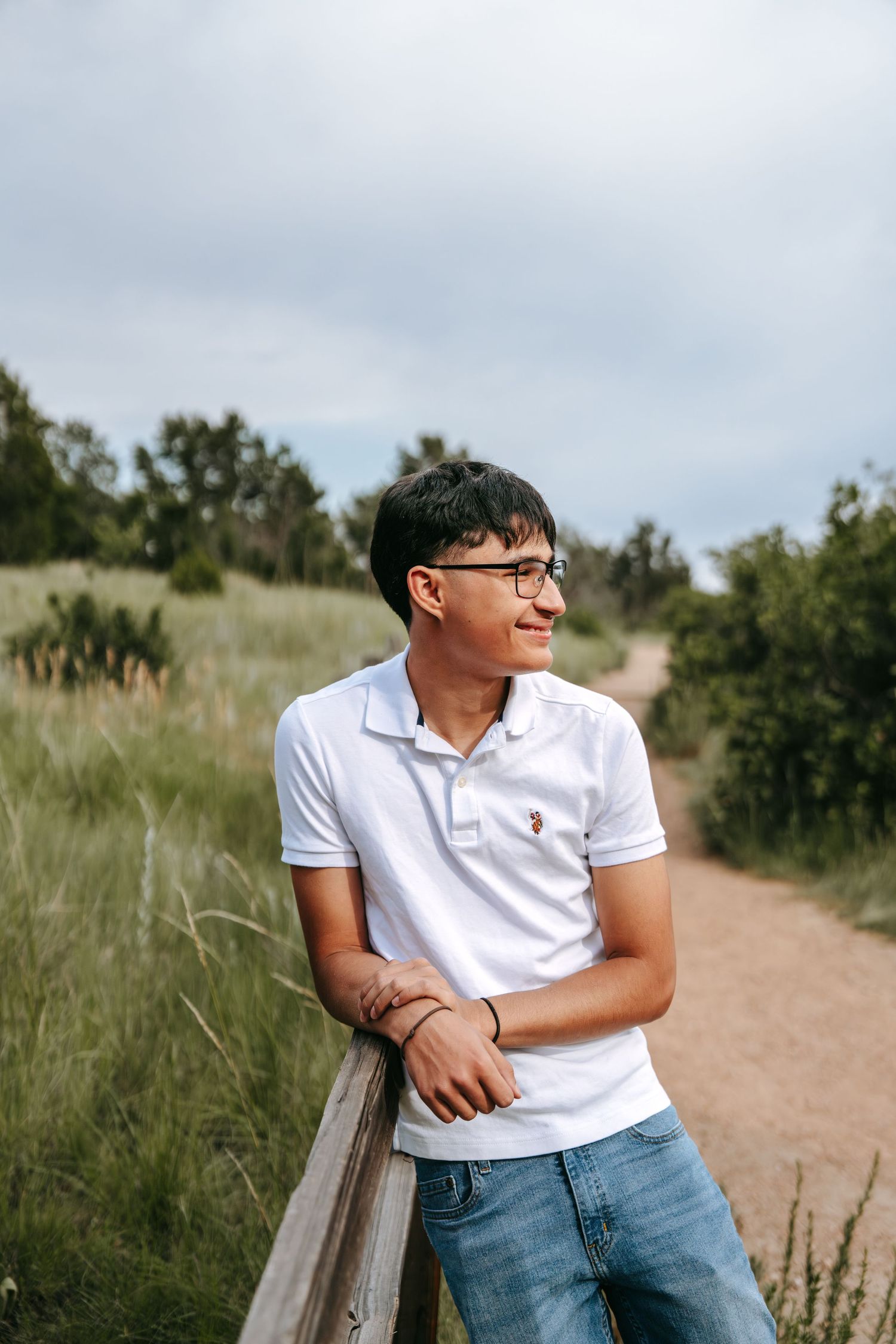 A young man in a white polo shirt leans on a wooden fence along a scenic nature trail.